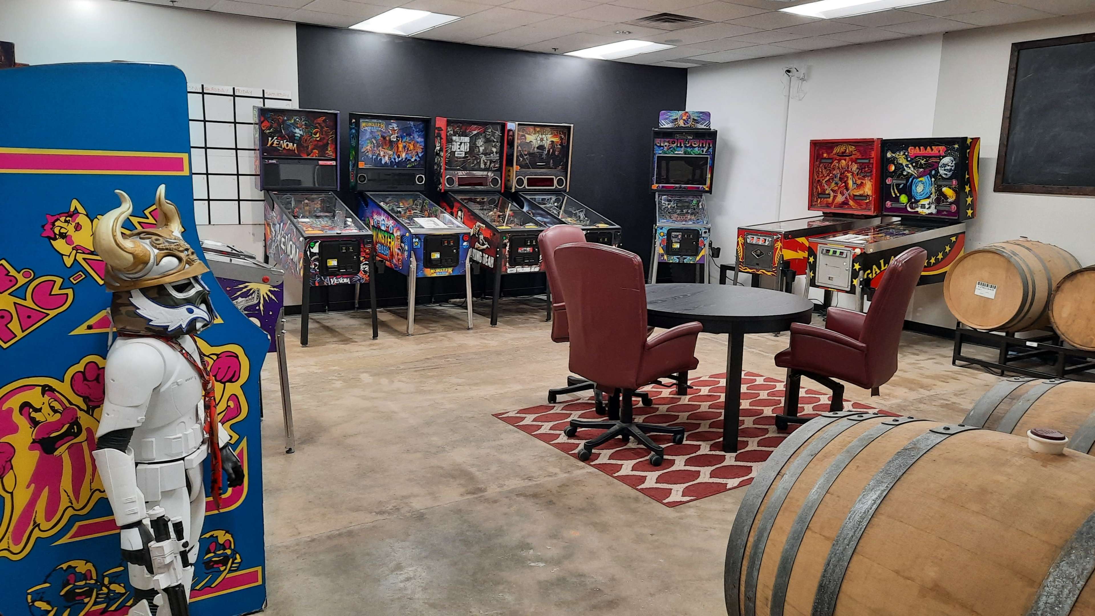 The image shows a room with several pinball machines, a round table with two chairs, and barrels in the corner.