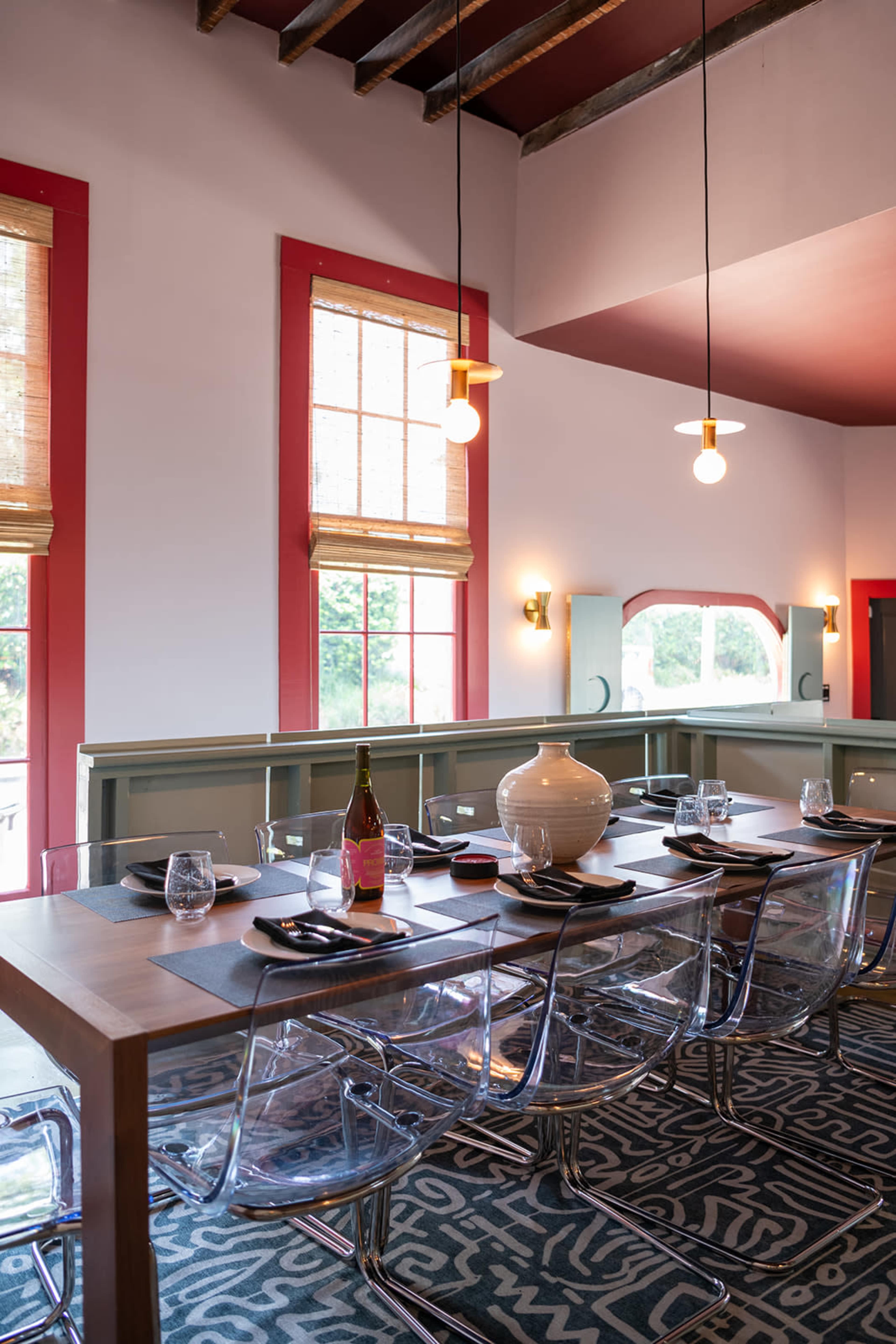 A long wooden dining table with clear plastic chairs is set against a backdrop of bright red windows and a patterned rug in a modern dining room.