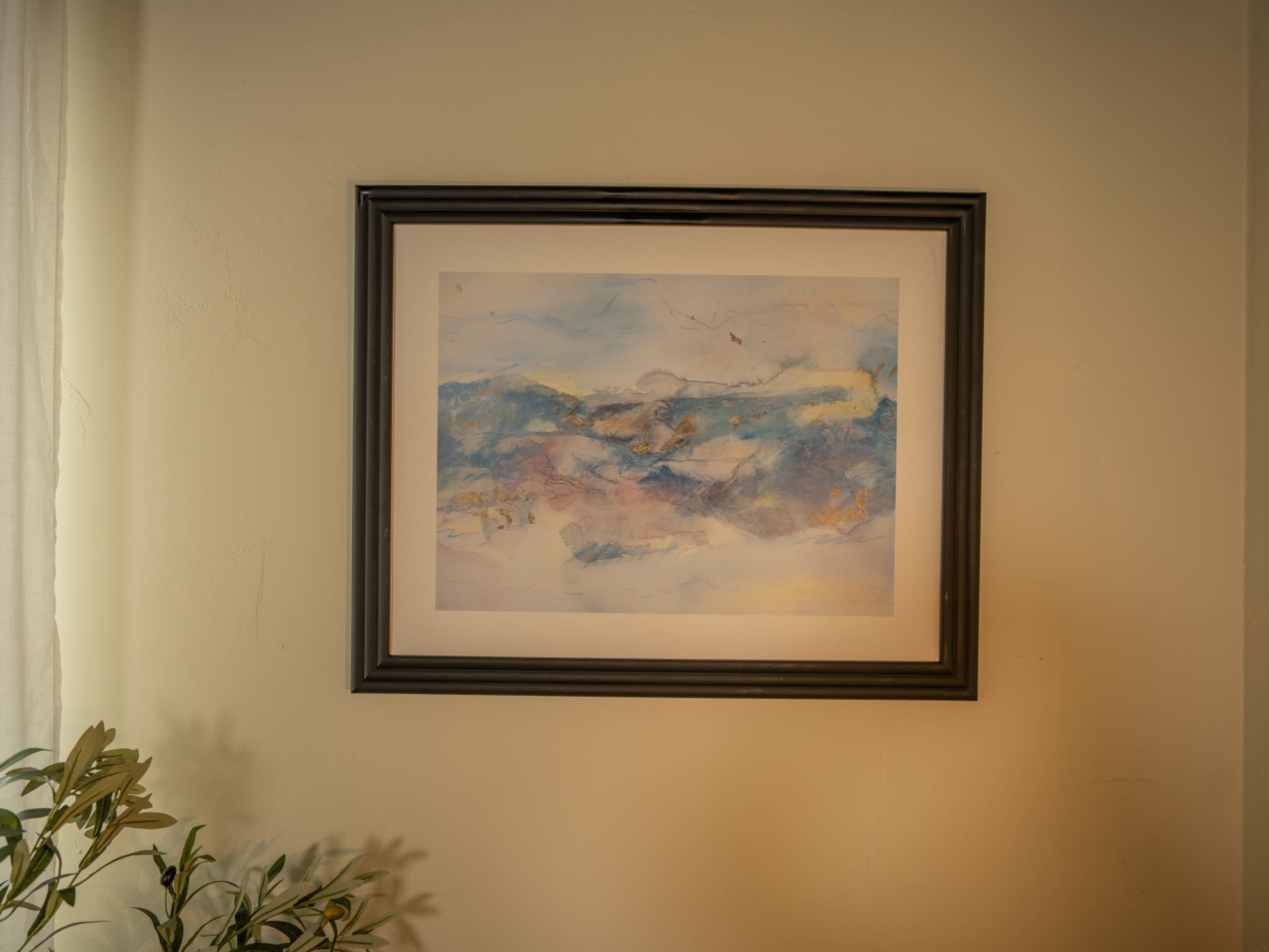 The image features a framed abstract watercolor painting hanging on a wall, with a plant partially visible in the foreground.