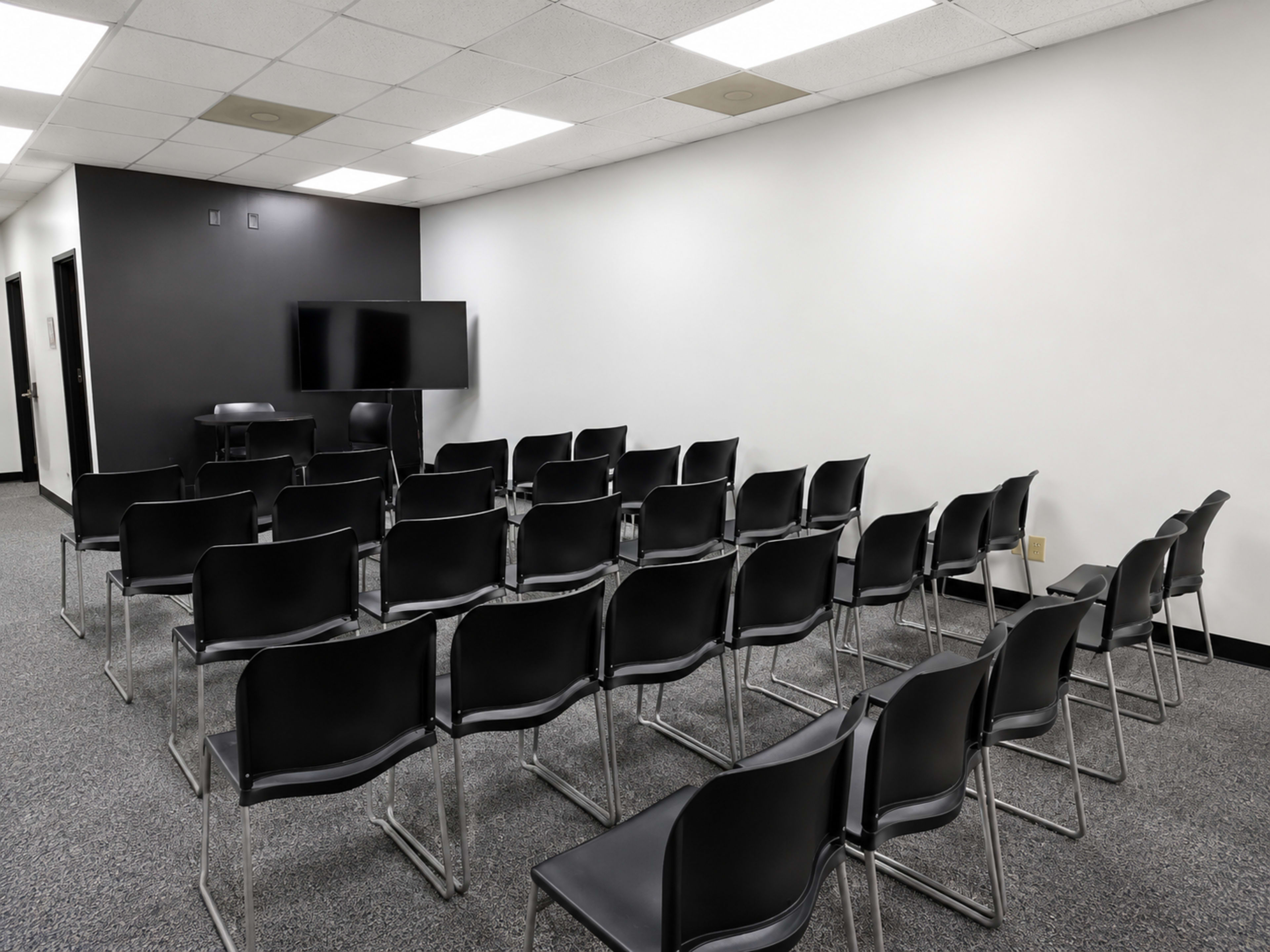 The image shows a conference room with rows of black chairs arranged in front of a blank television screen and a dark wall.