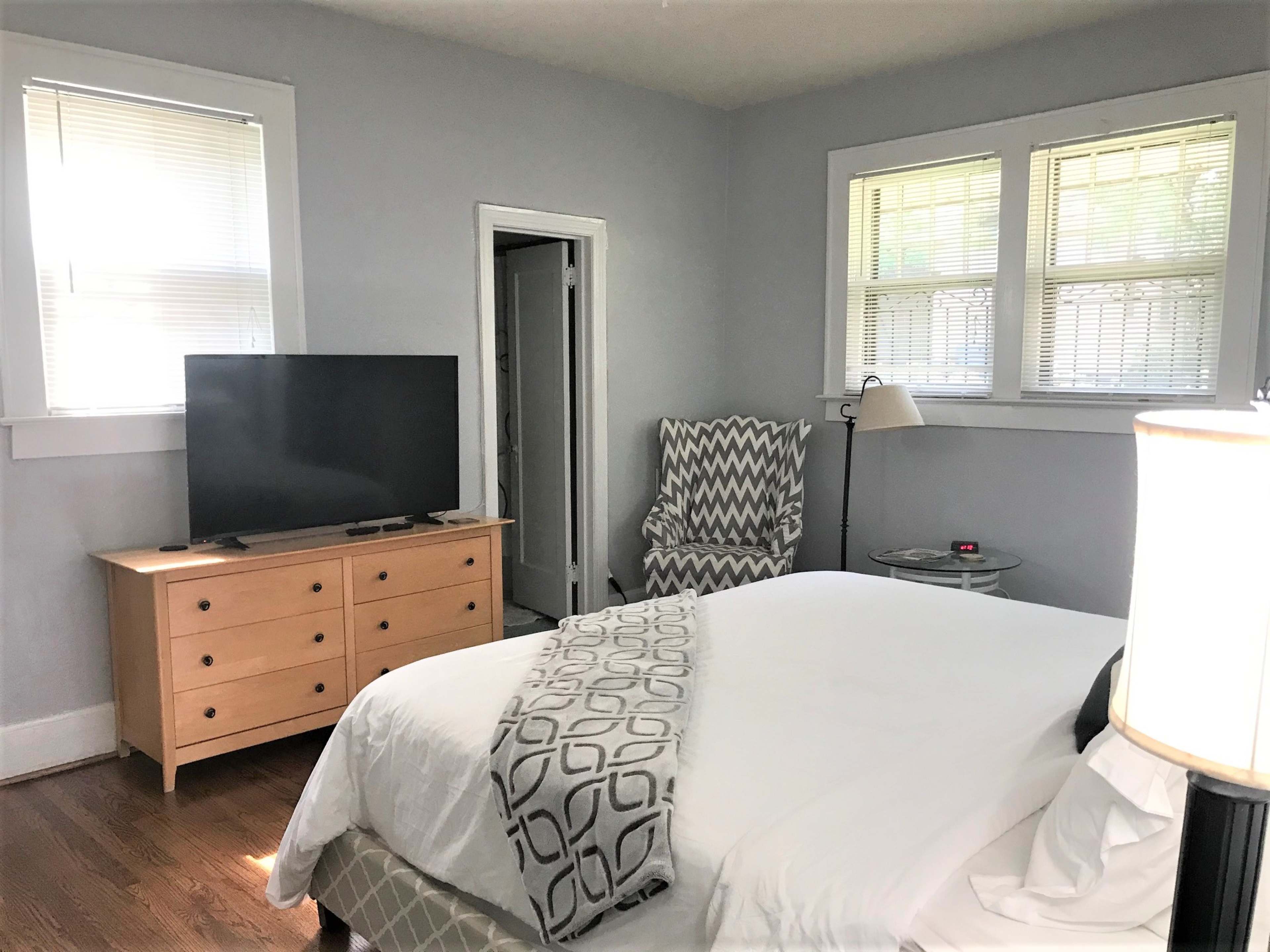 The image shows a tidy bedroom with a bed, a television on a wooden dresser, and a chair next to a lamp near the window.