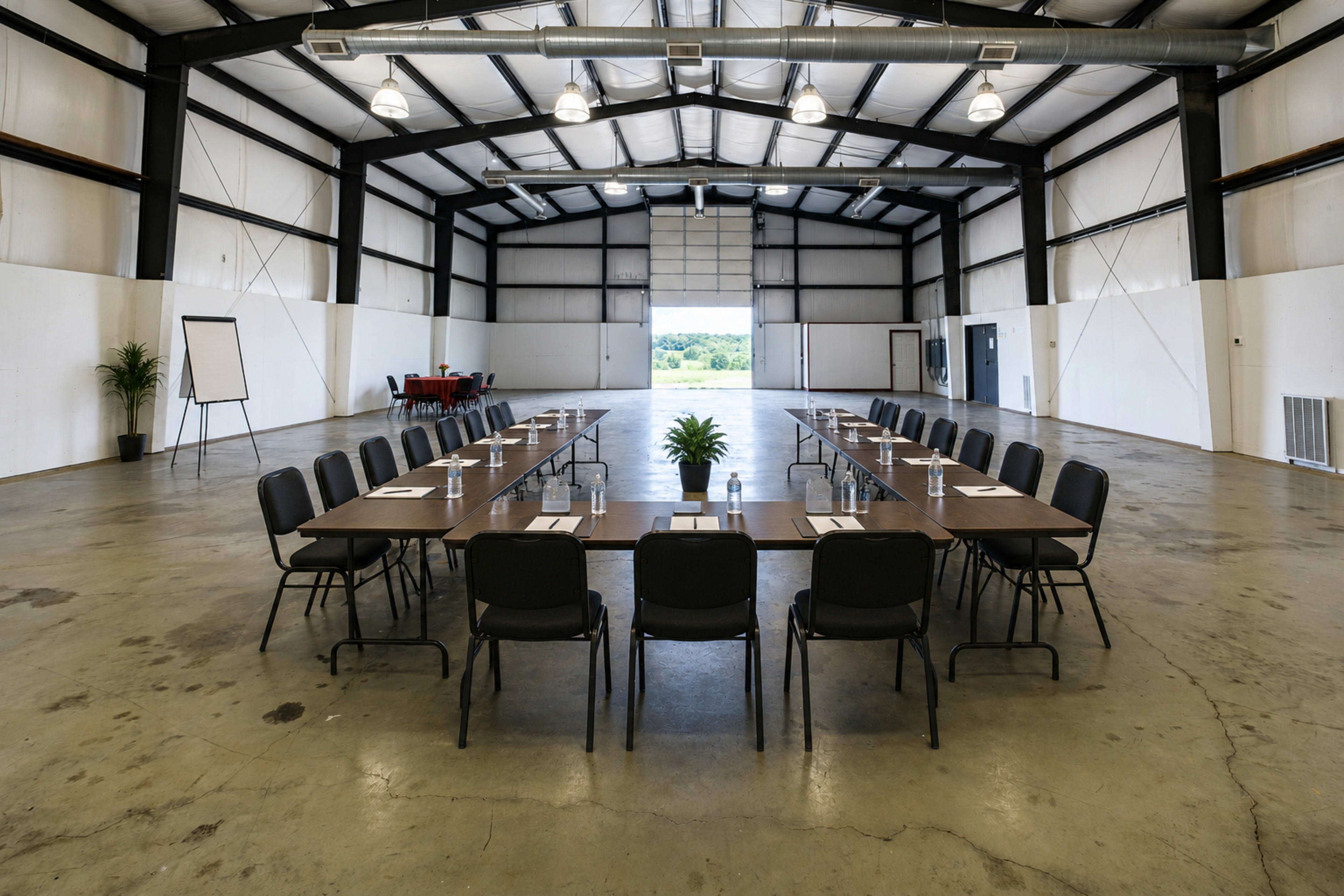A spacious meeting room features a long table arranged with chairs and materials, set within a large industrial space with an open door leading to an outdoor view.