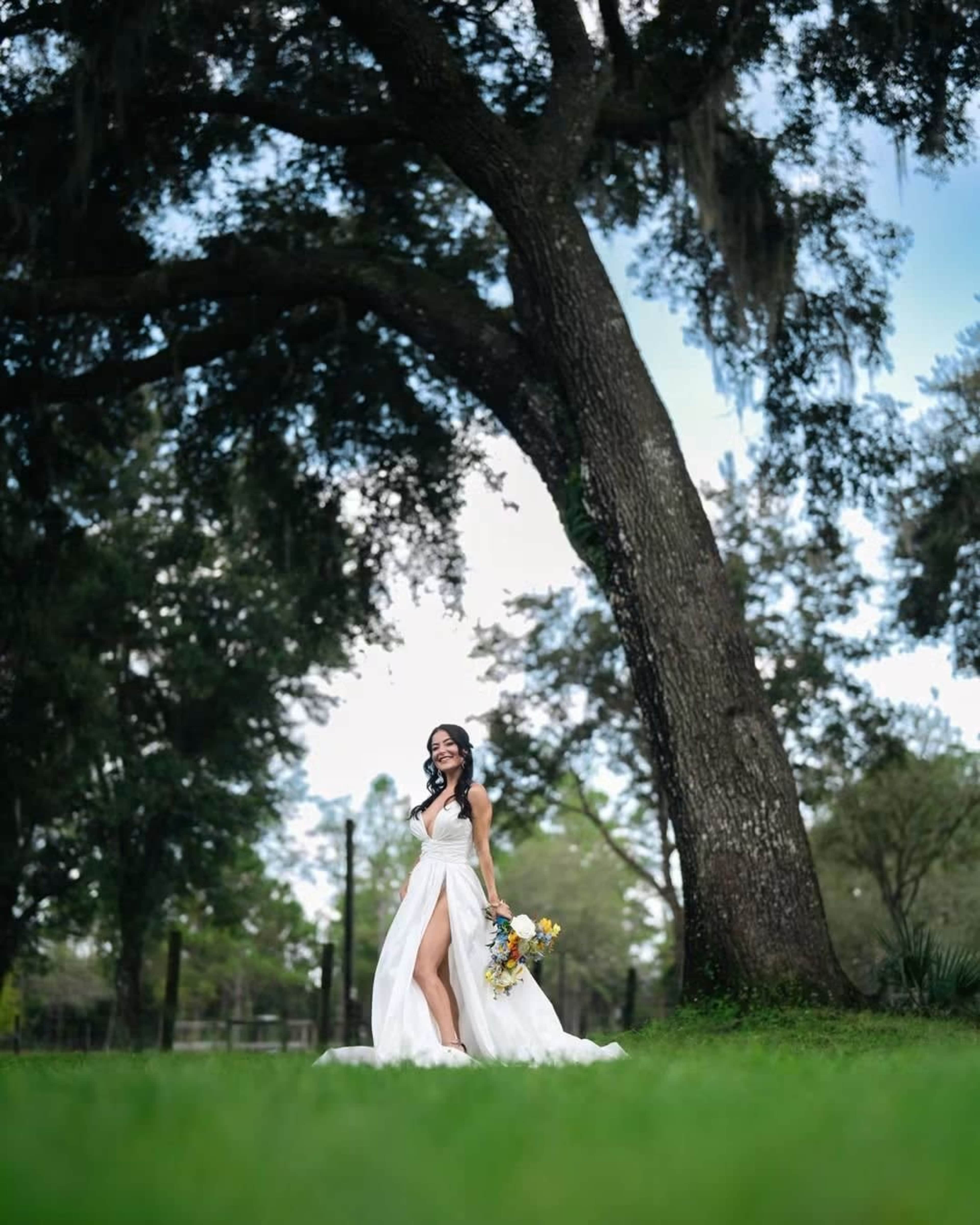A bride in a white gown stands in a grassy area under a large tree, holding a bouquet of flowers.