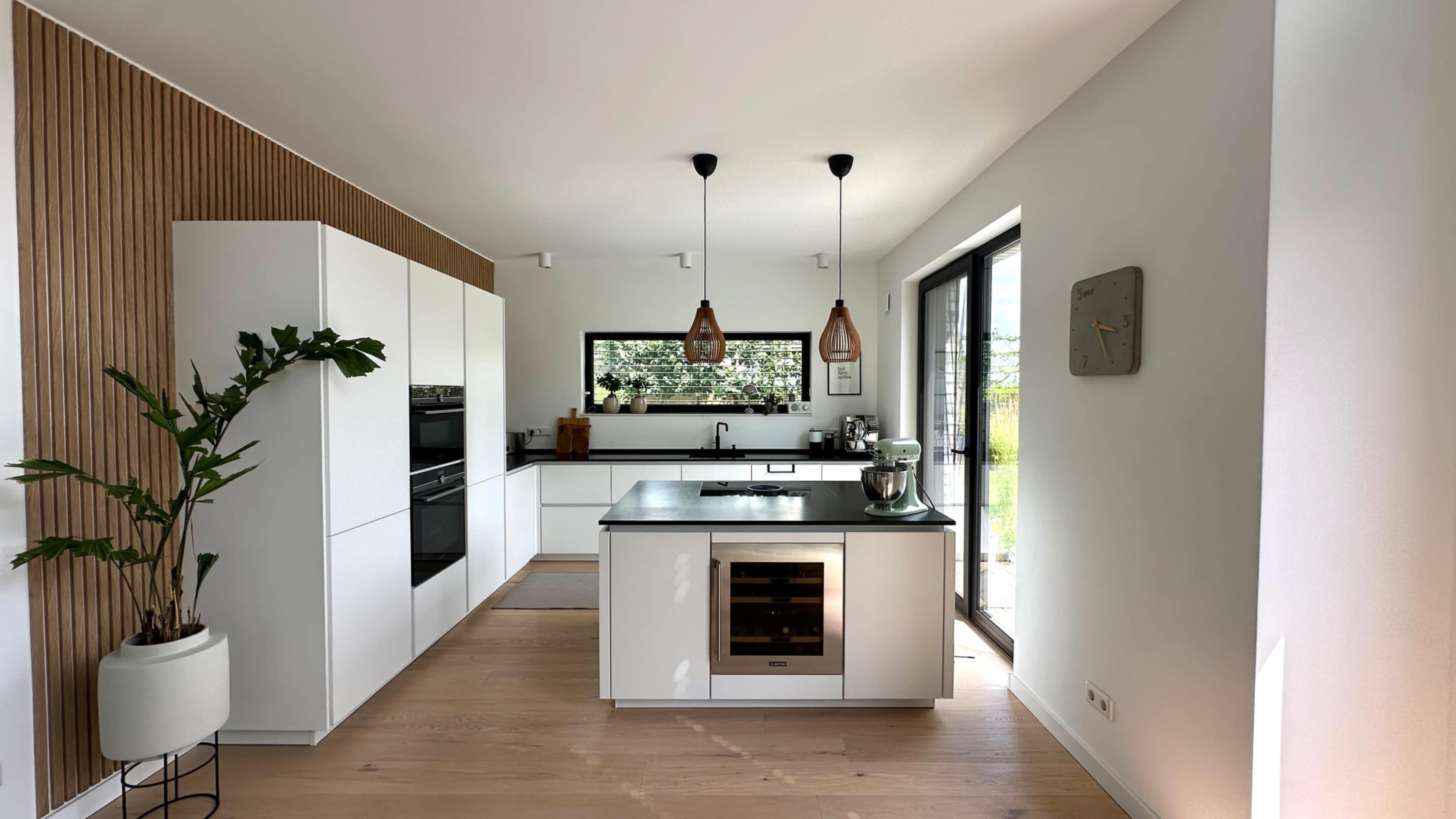 The image shows a modern kitchen with white cabinetry, a central island, and large windows that provide natural light.