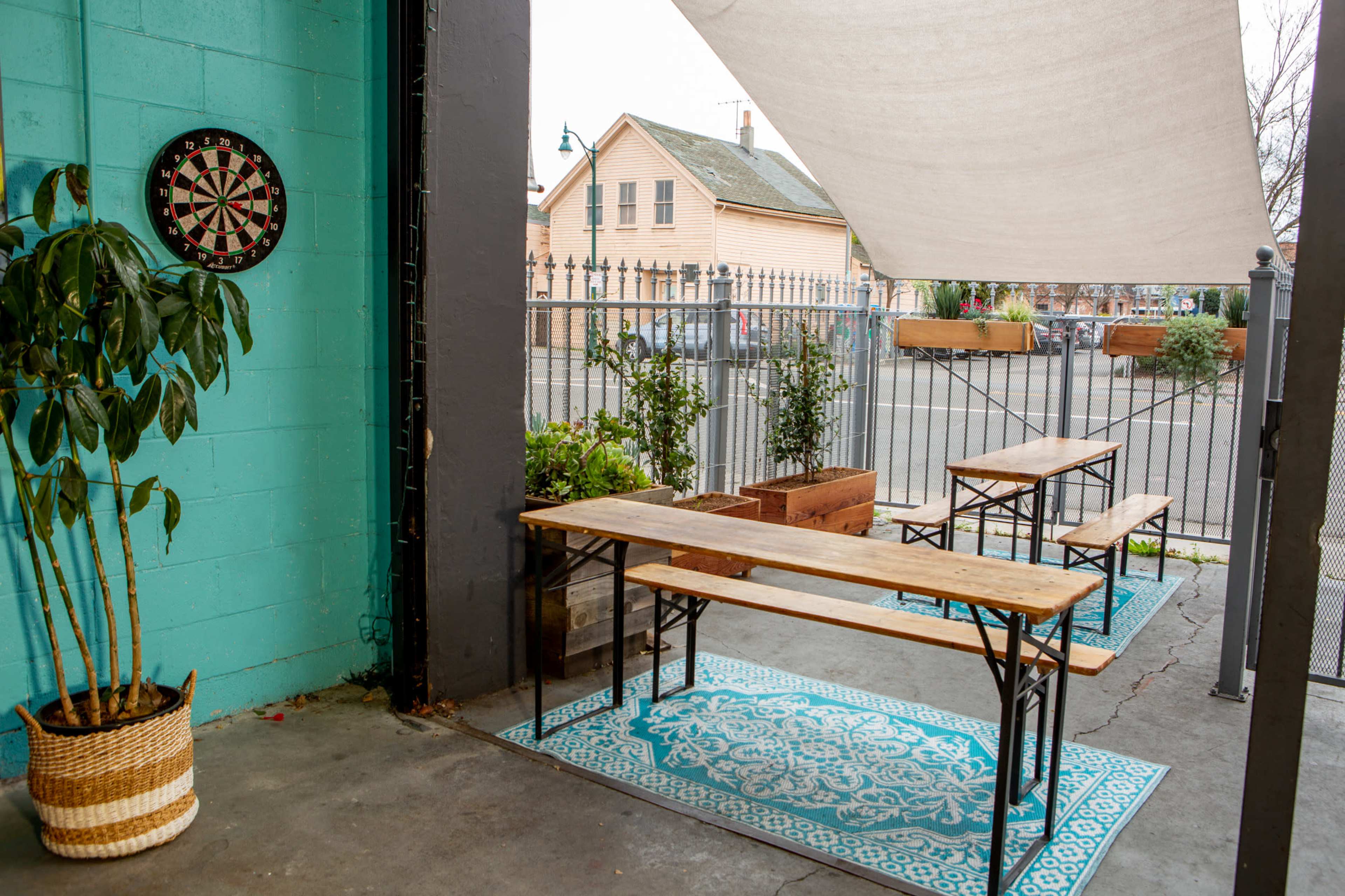 The image shows an outdoor seating area with wooden benches, a dartboard on a turquoise wall, and potted plants, under a canopy.