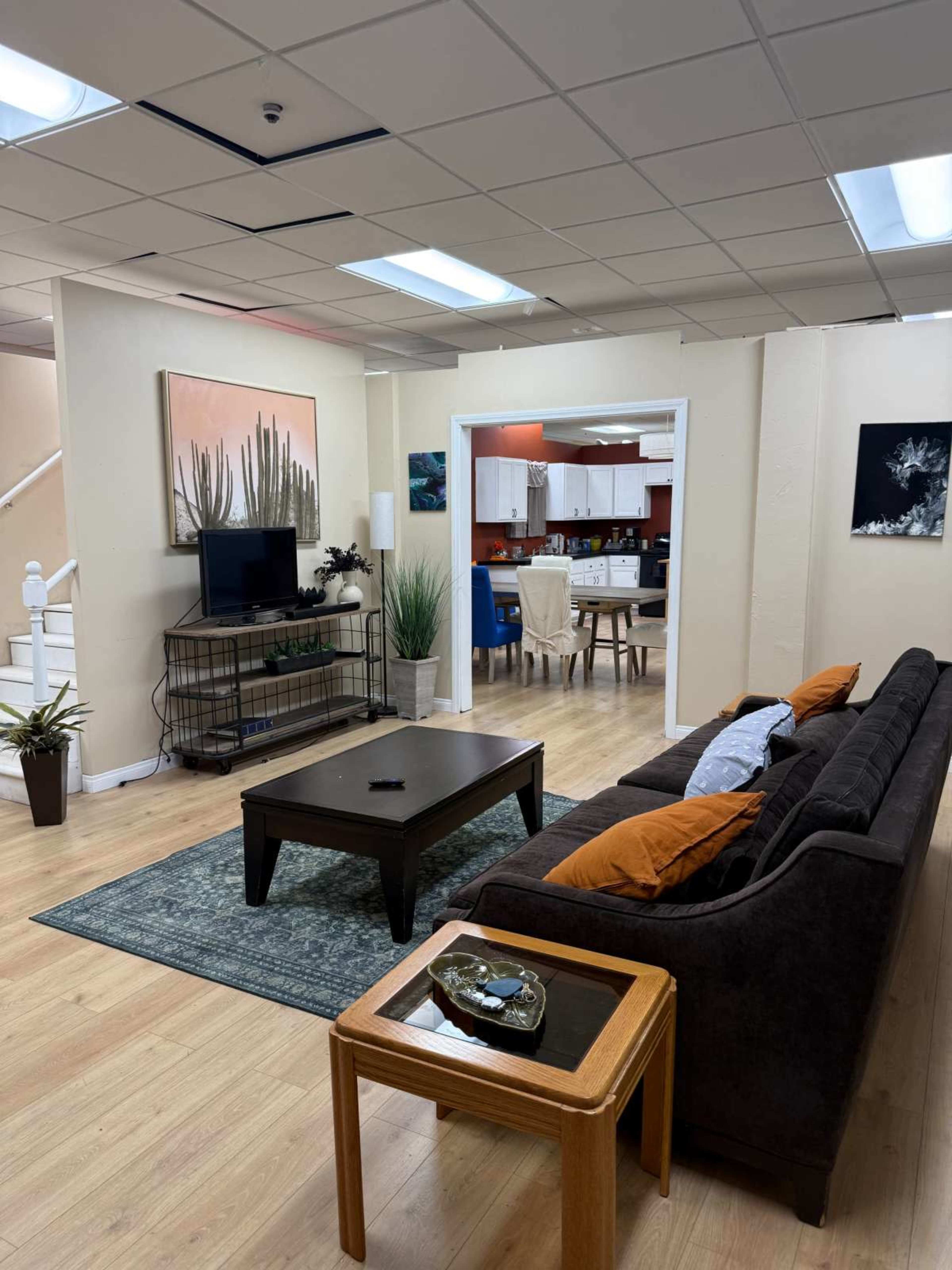 The image shows a living room with a dark sofa, a coffee table, and a view into a kitchen area, featuring a larger rug and decorative plants.