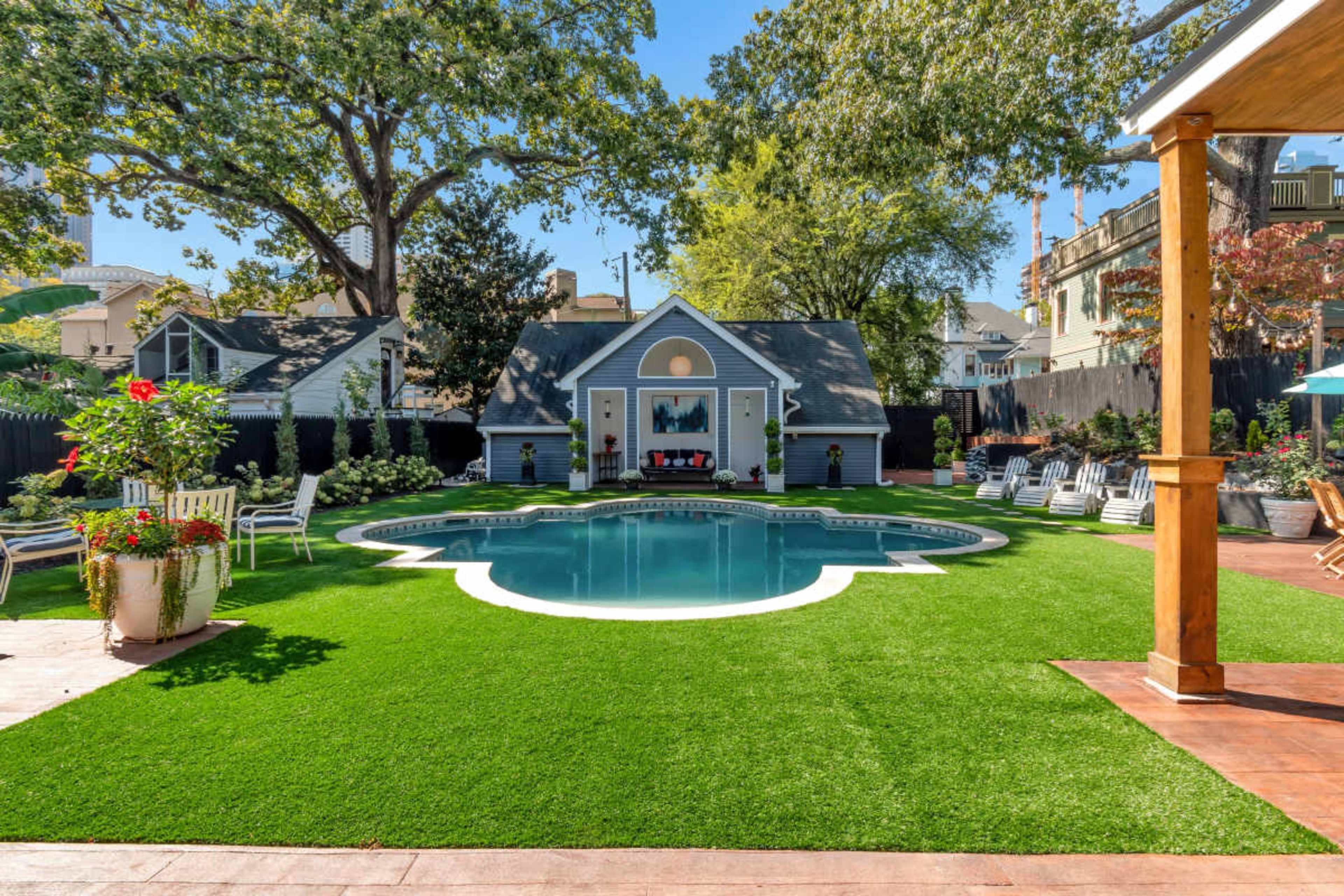 The image shows a backyard pool area with a neatly manicured lawn, surrounded by lounge chairs, colorful flowers, and a small building in the background.