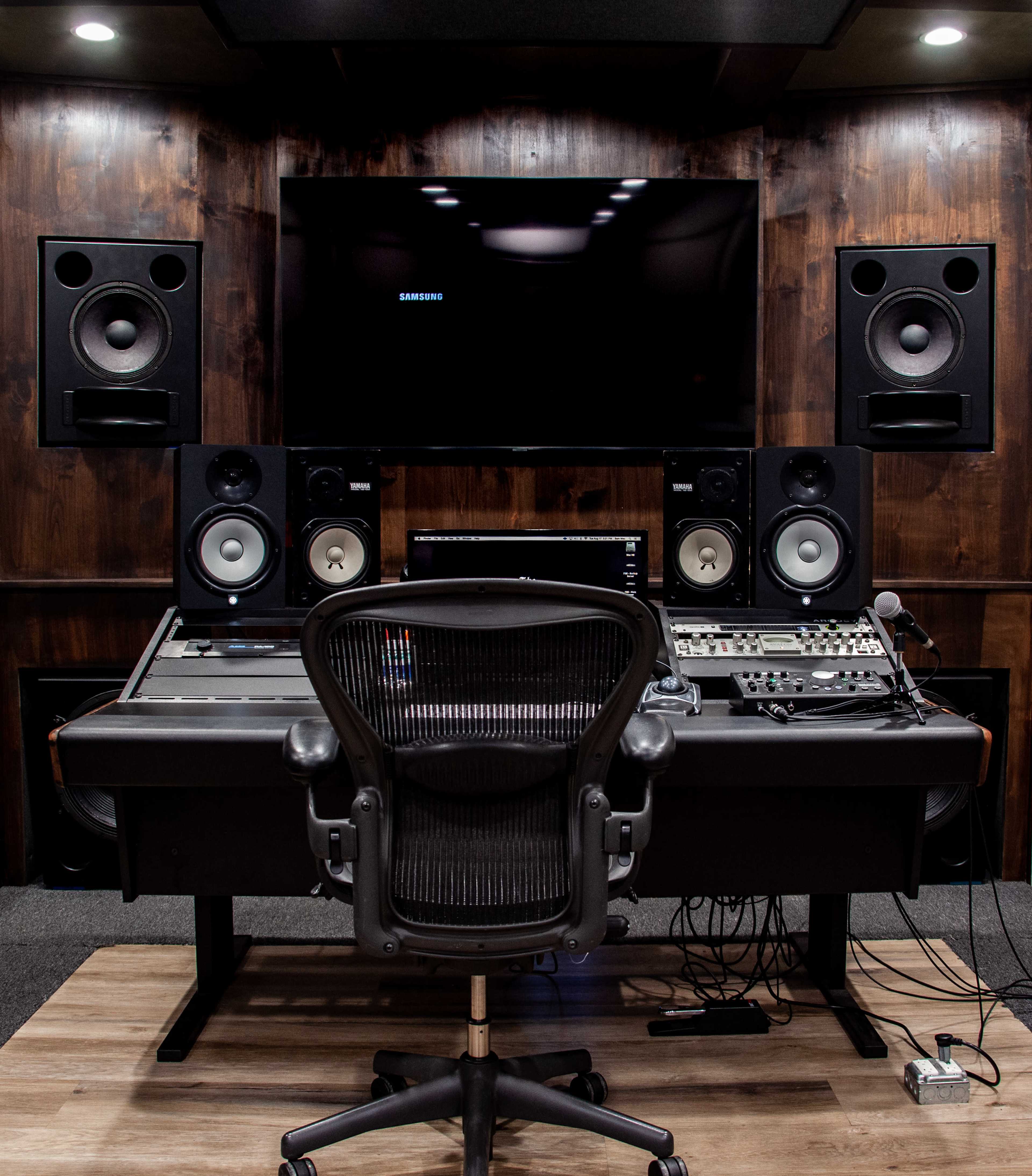 A recording studio features a sound mixing desk, speakers, and a large screen, all set against a dark wooden wall.