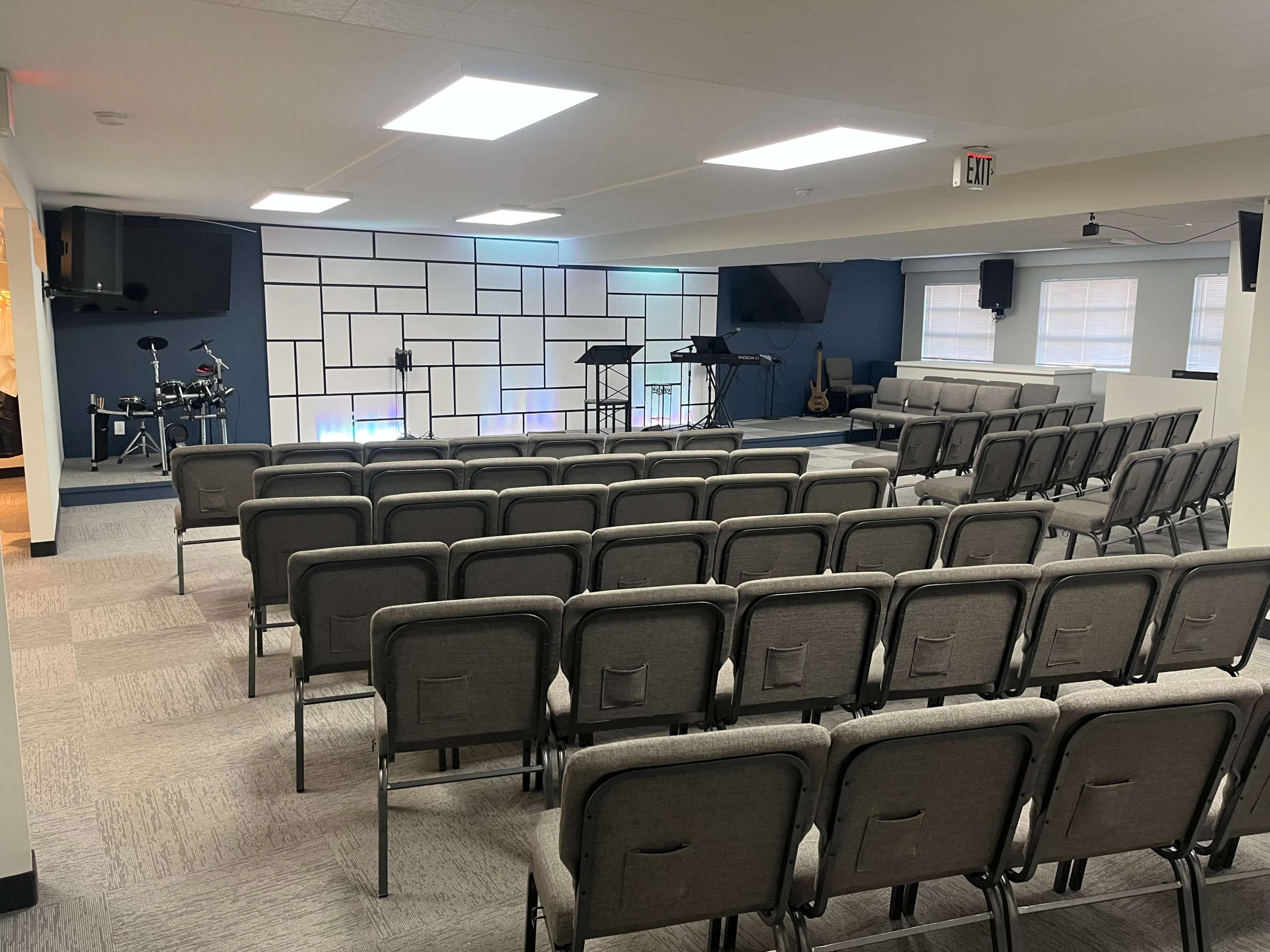 A spacious room arranged with rows of gray chairs facing a stage, which has musical instruments and a backdrop featuring geometric wall designs.