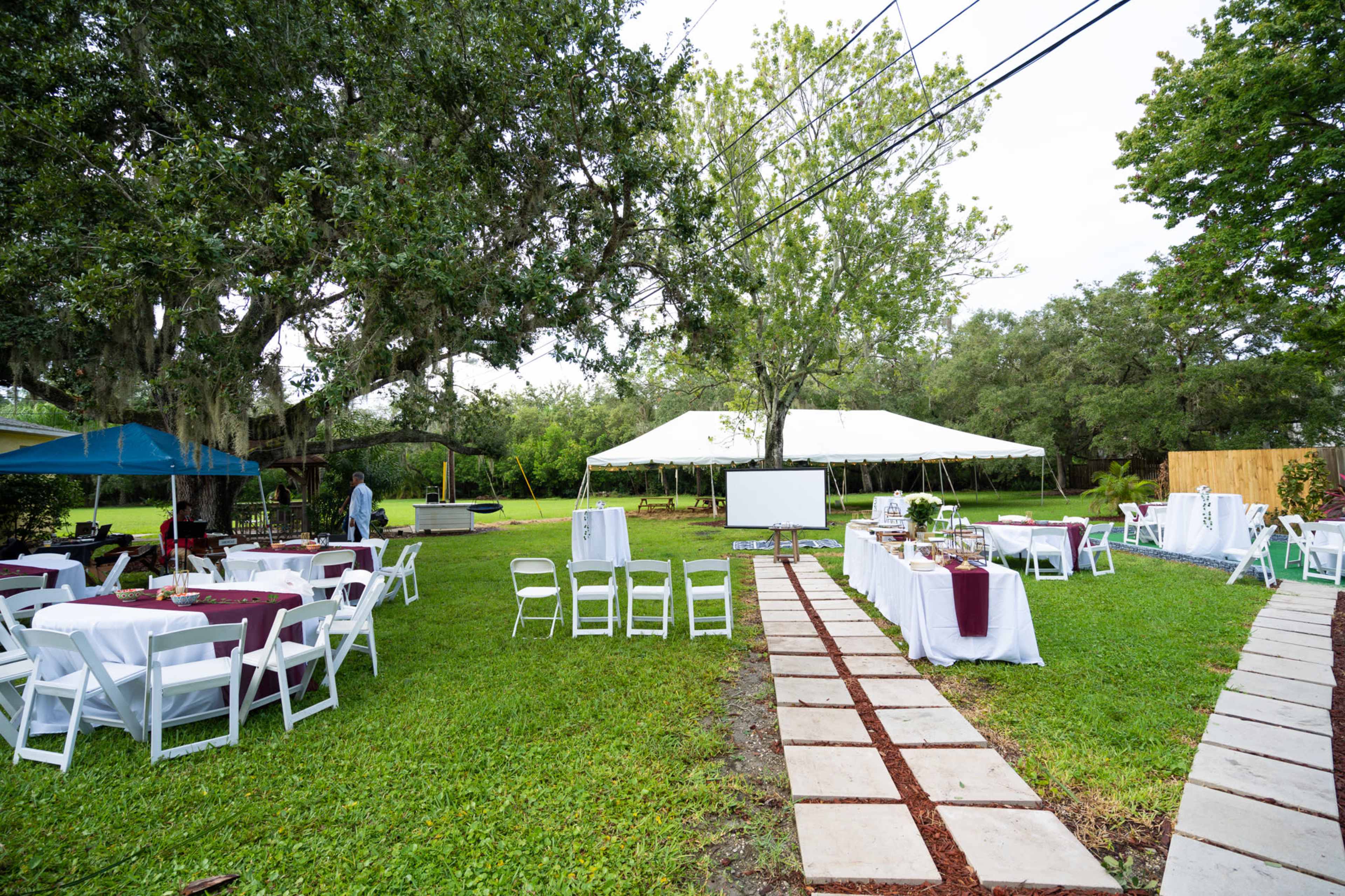 The outdoor event space features white tables with maroon tablecloths, a large tent, and a paved pathway surrounded by greenery.
