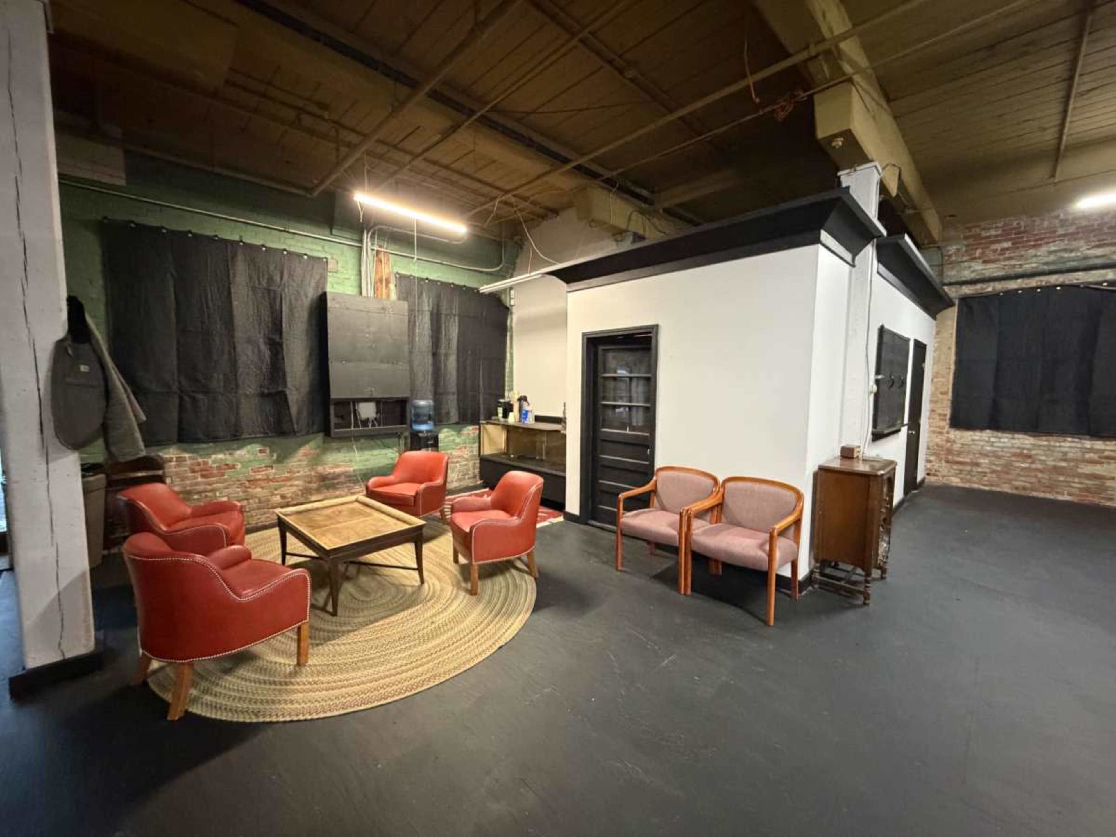 The image shows a spacious room featuring a circular rug, a set of four red chairs arranged around a wooden table, and a small kitchenette area in the background.