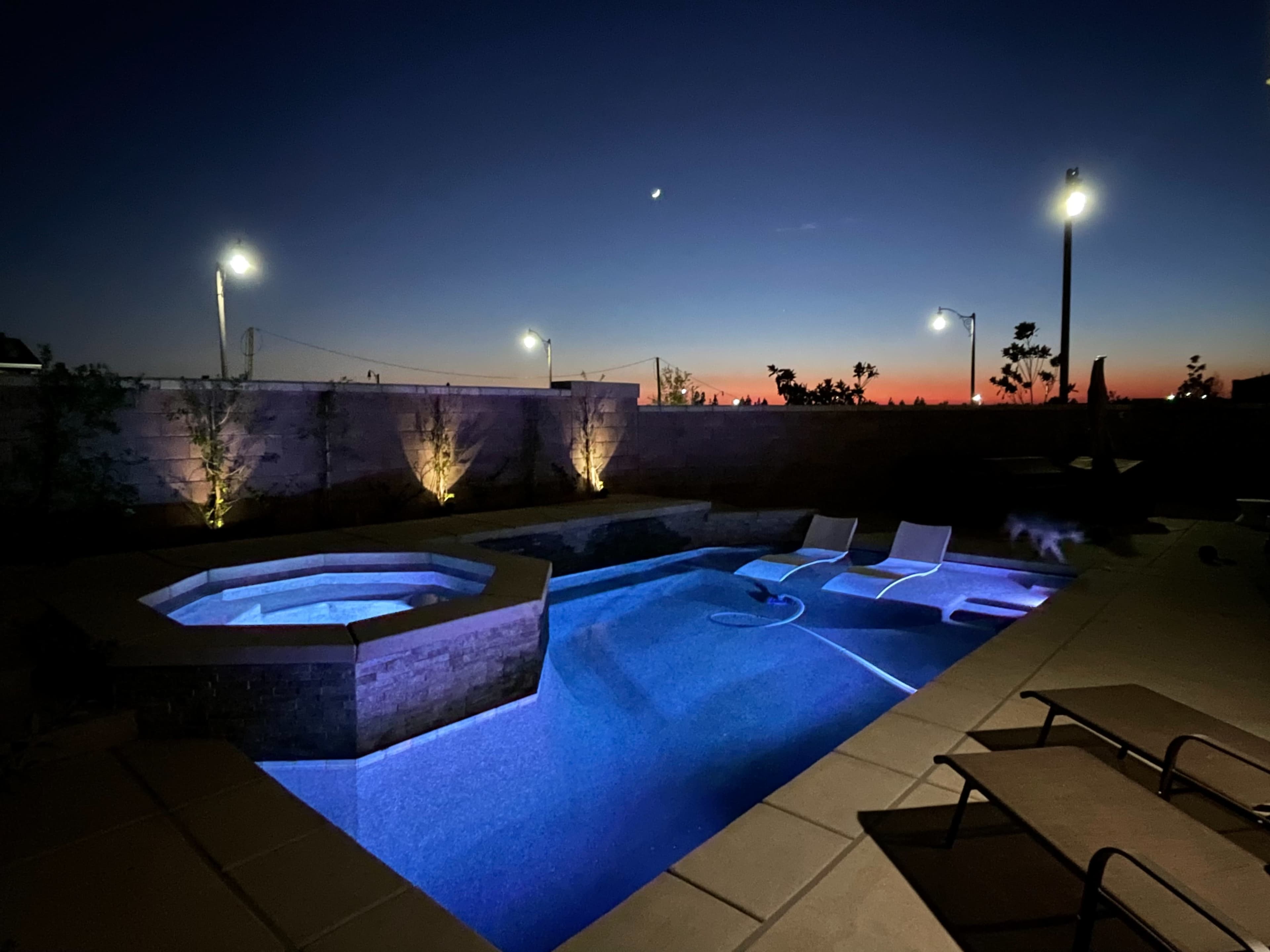 A backyard pool with a hot tub and lounge chairs is illuminated by blue lighting under a twilight sky.