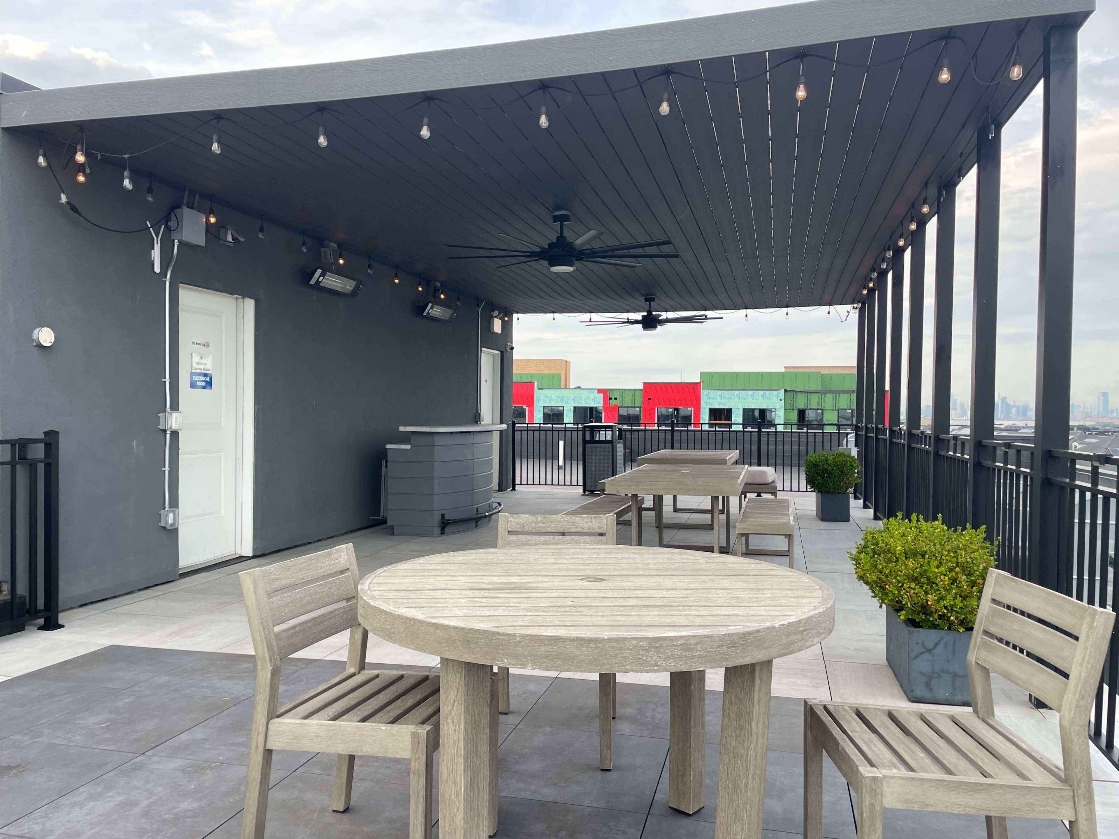 A rooftop terrace with wooden tables and chairs under a covered area, string lights overhead, and a view of buildings in the background.