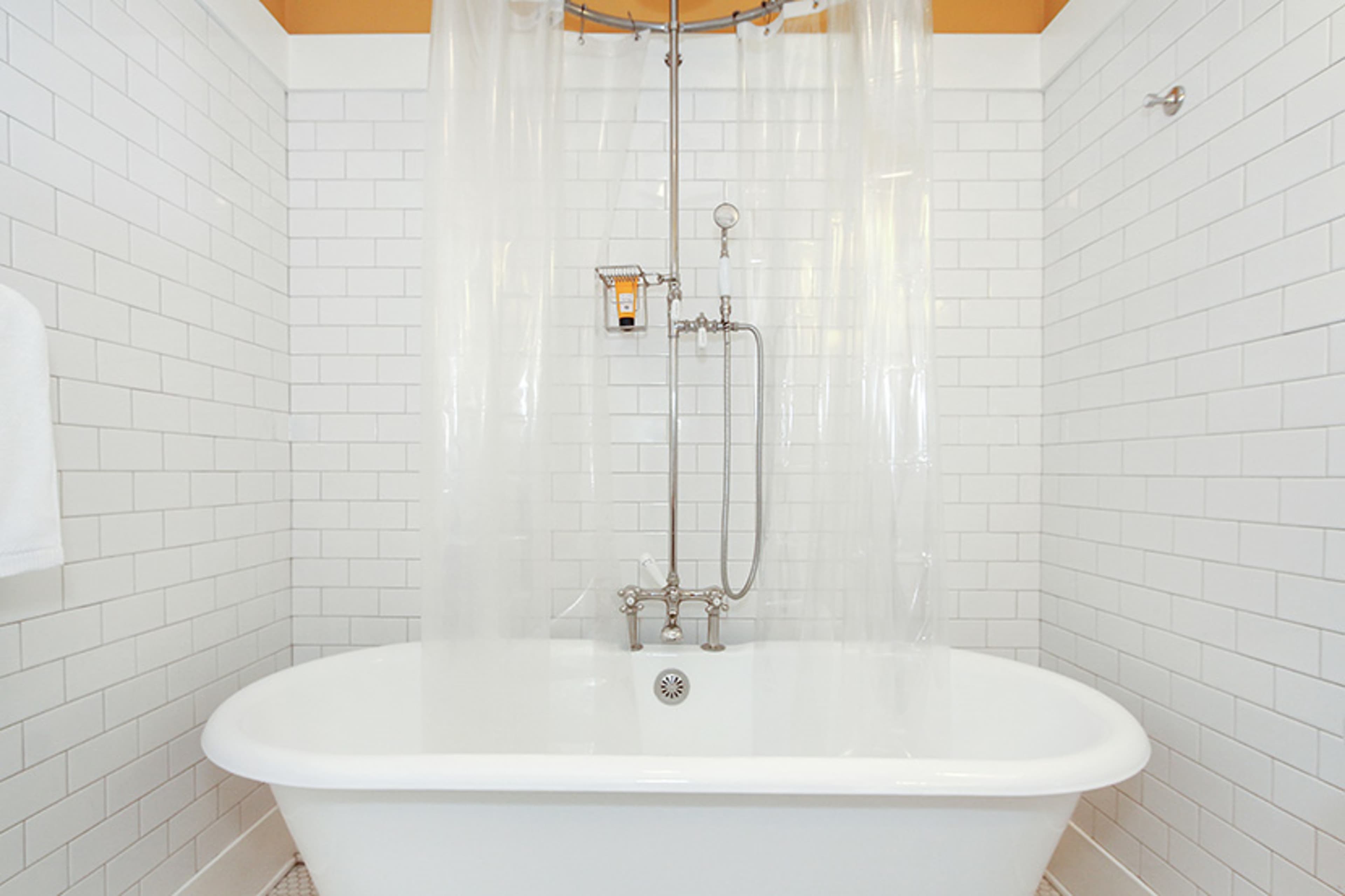 A freestanding bathtub is surrounded by white tiled walls and a clear shower curtain.