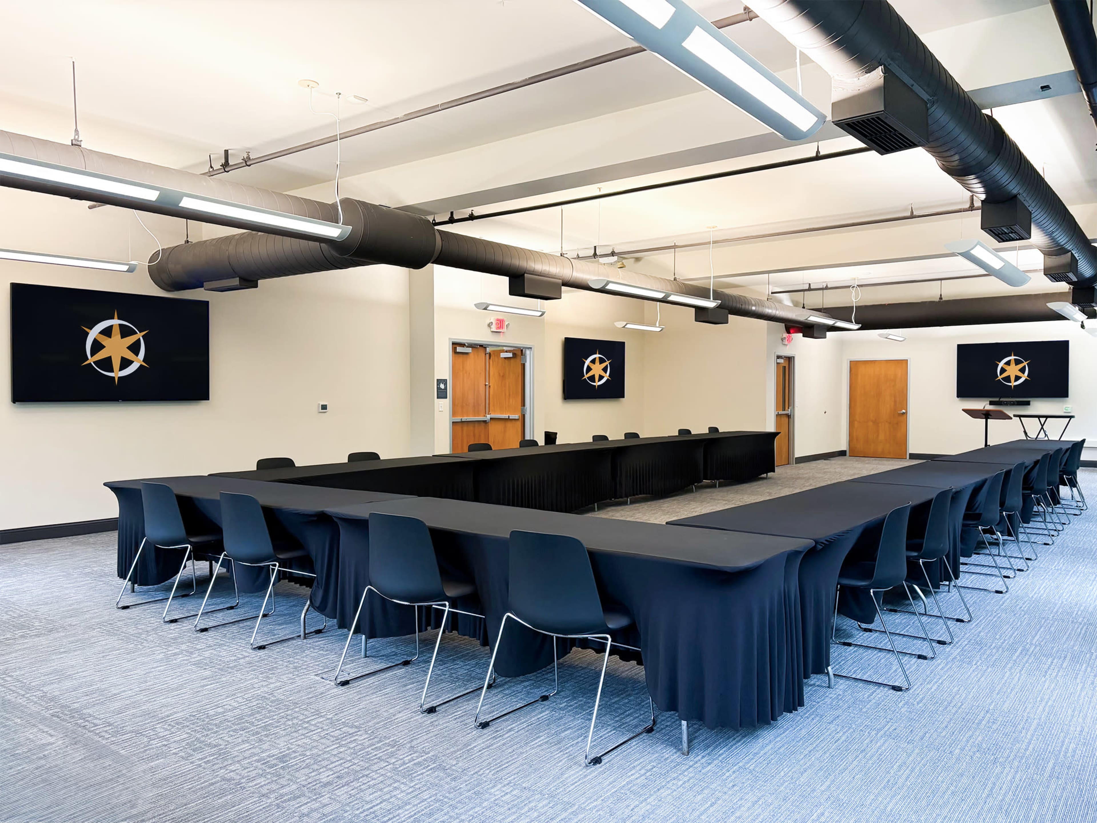 The image shows a modern conference room arranged with a large rectangular table surrounded by chairs, with screens displaying a logo on the walls.