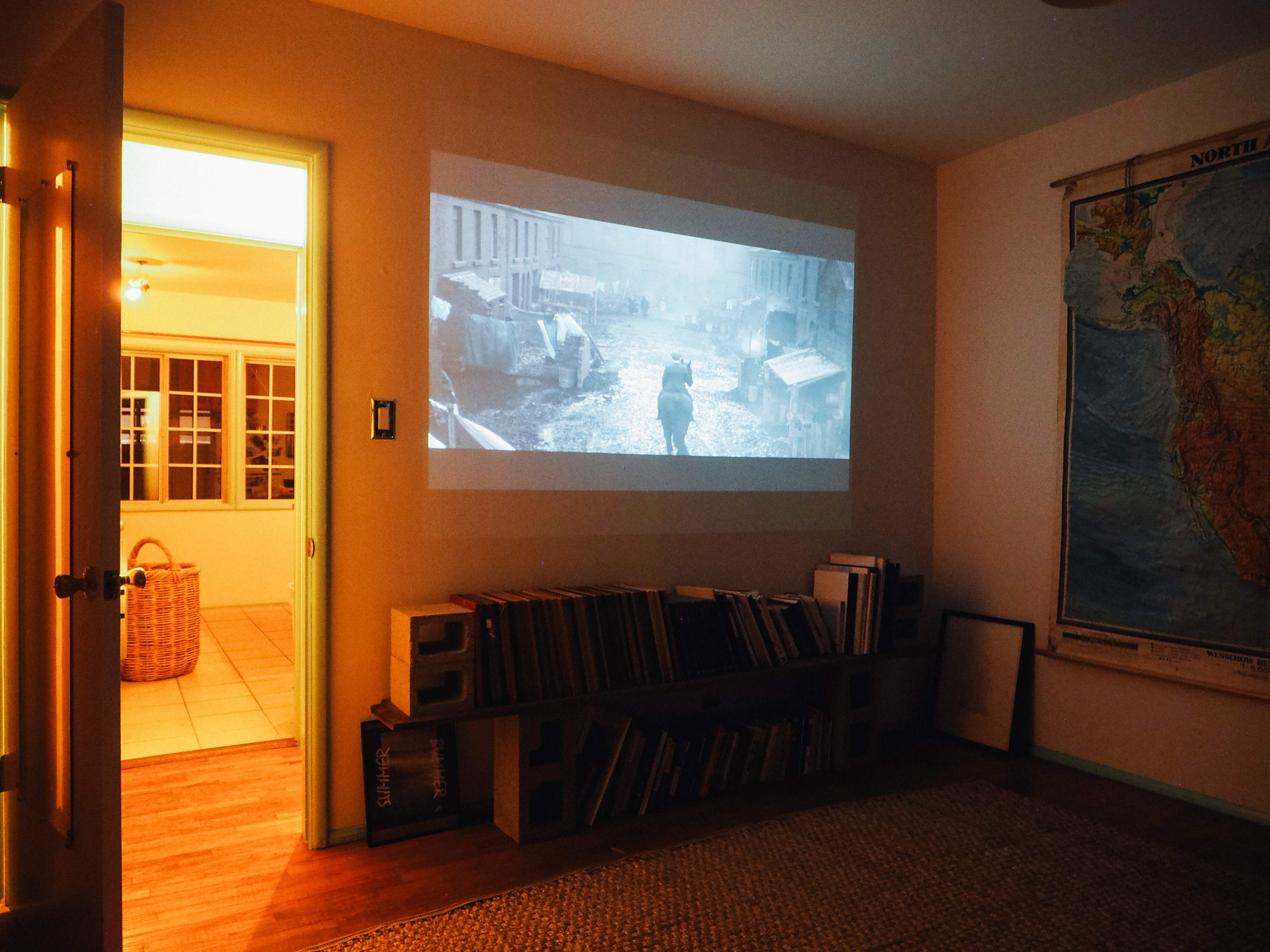 A black-and-white film is projected onto a wall in a dimly lit room with a doorway leading to a brighter space.