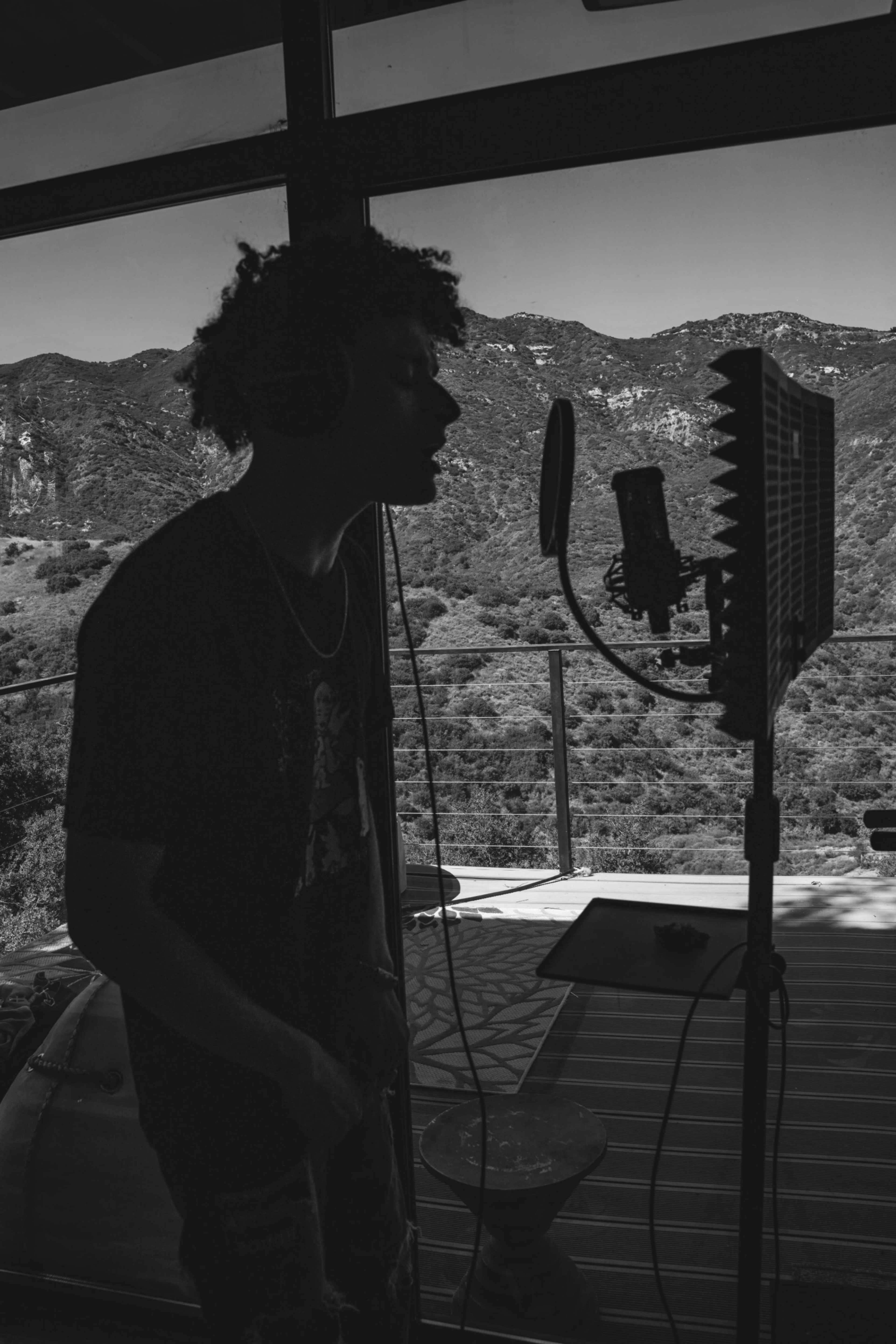 A person stands in profile, recording vocals into a microphone in a room with large windows overlooking a mountainous landscape.