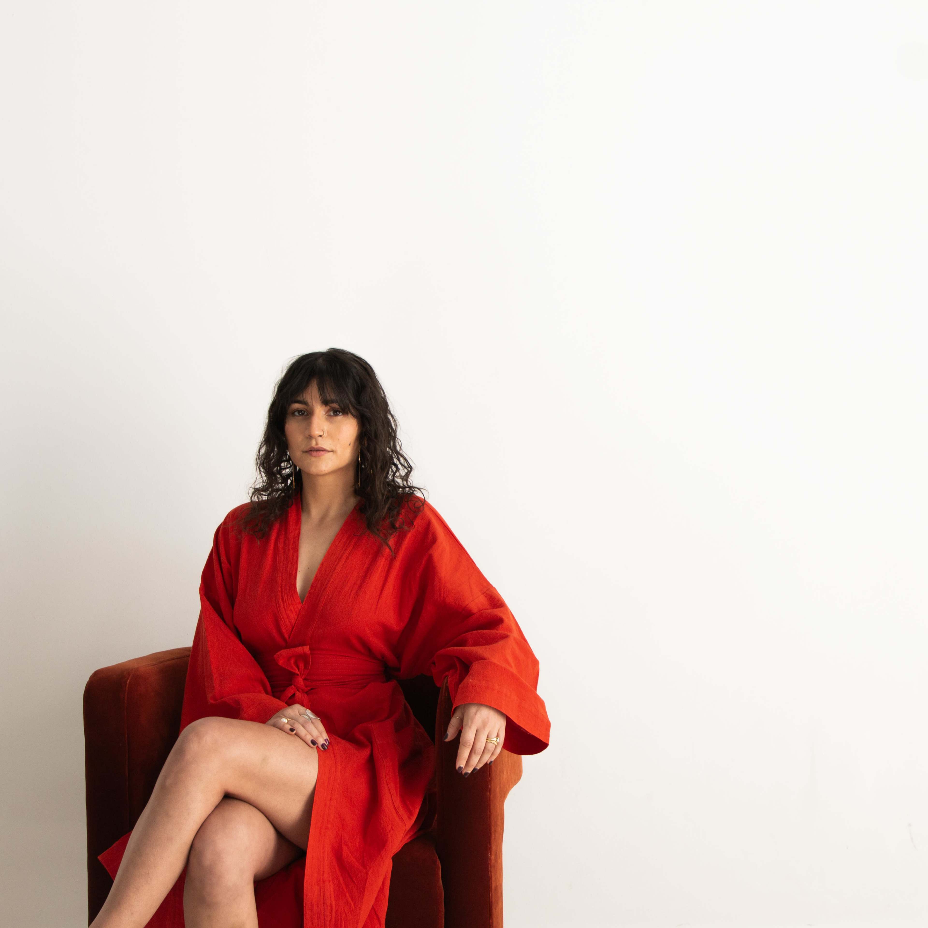 A woman in a red robe sits on a red armchair against a white background.