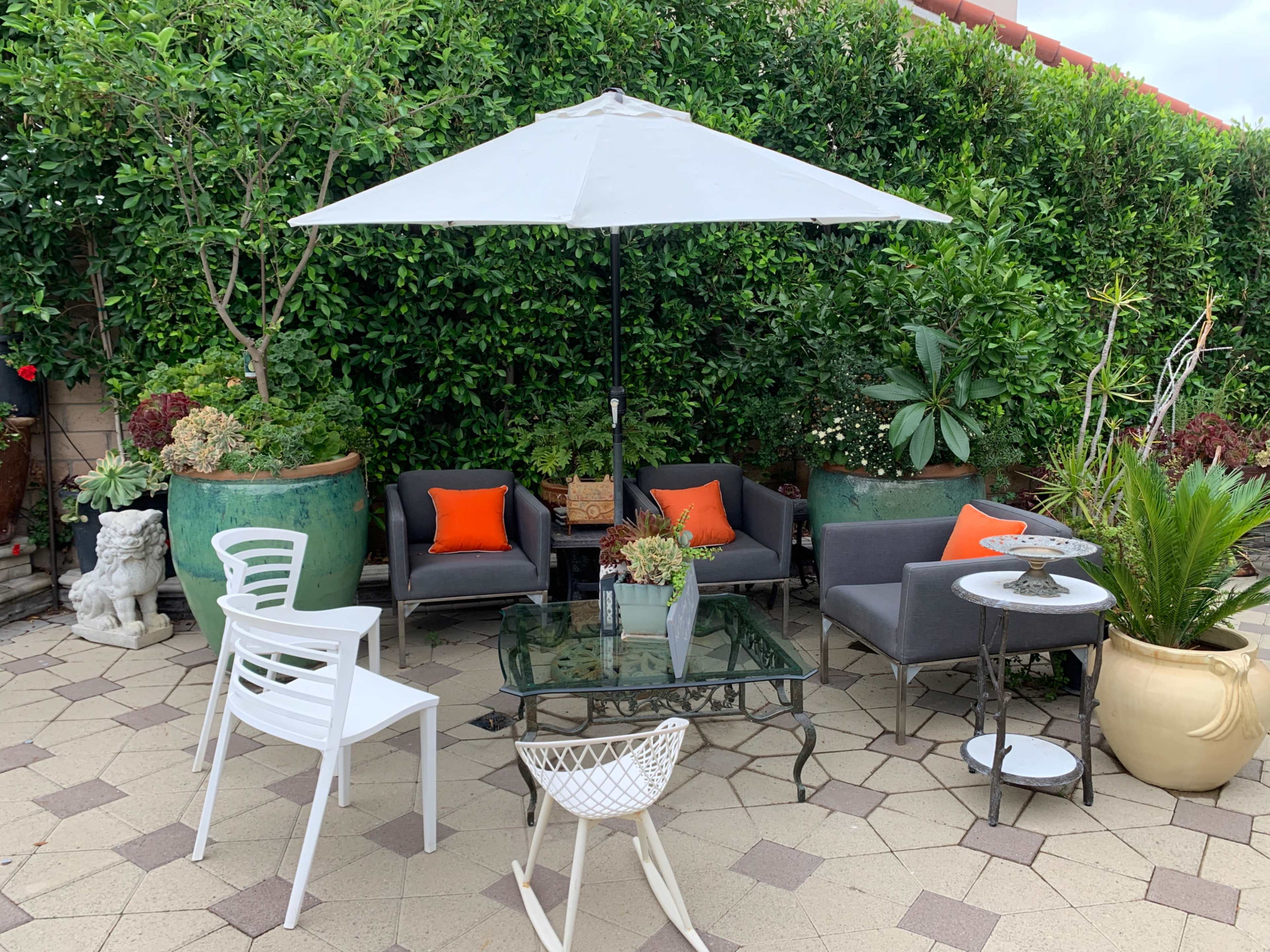 A patio area with gray seating, orange cushions, a glass table, and a large umbrella, surrounded by green plants and decorative pots.