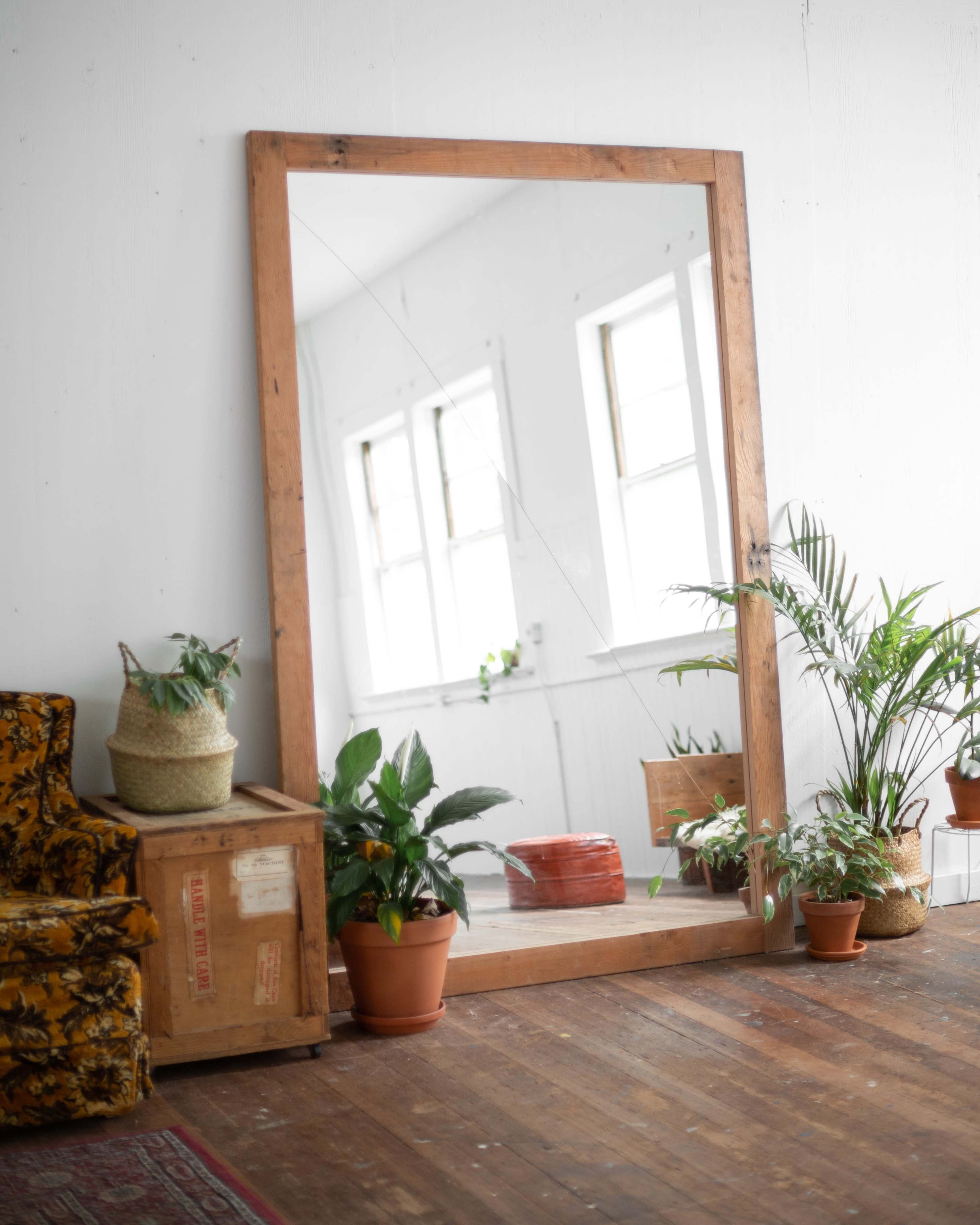 A large wooden-framed mirror stands against a wall next to a vintage sofa and several potted plants in a bright room.