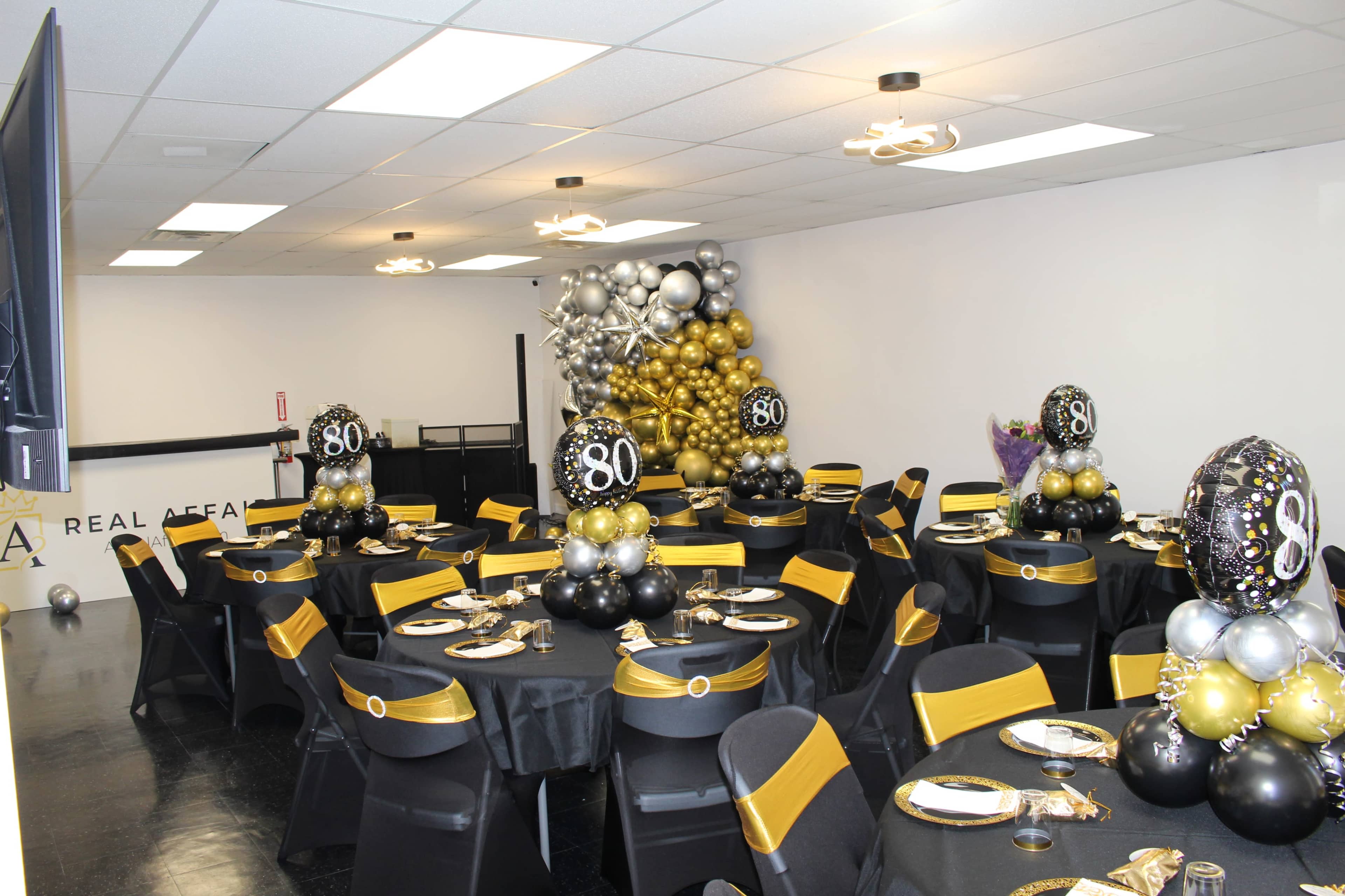 A decorated banquet room features black and gold table settings with balloon centerpieces celebrating an 80th birthday.