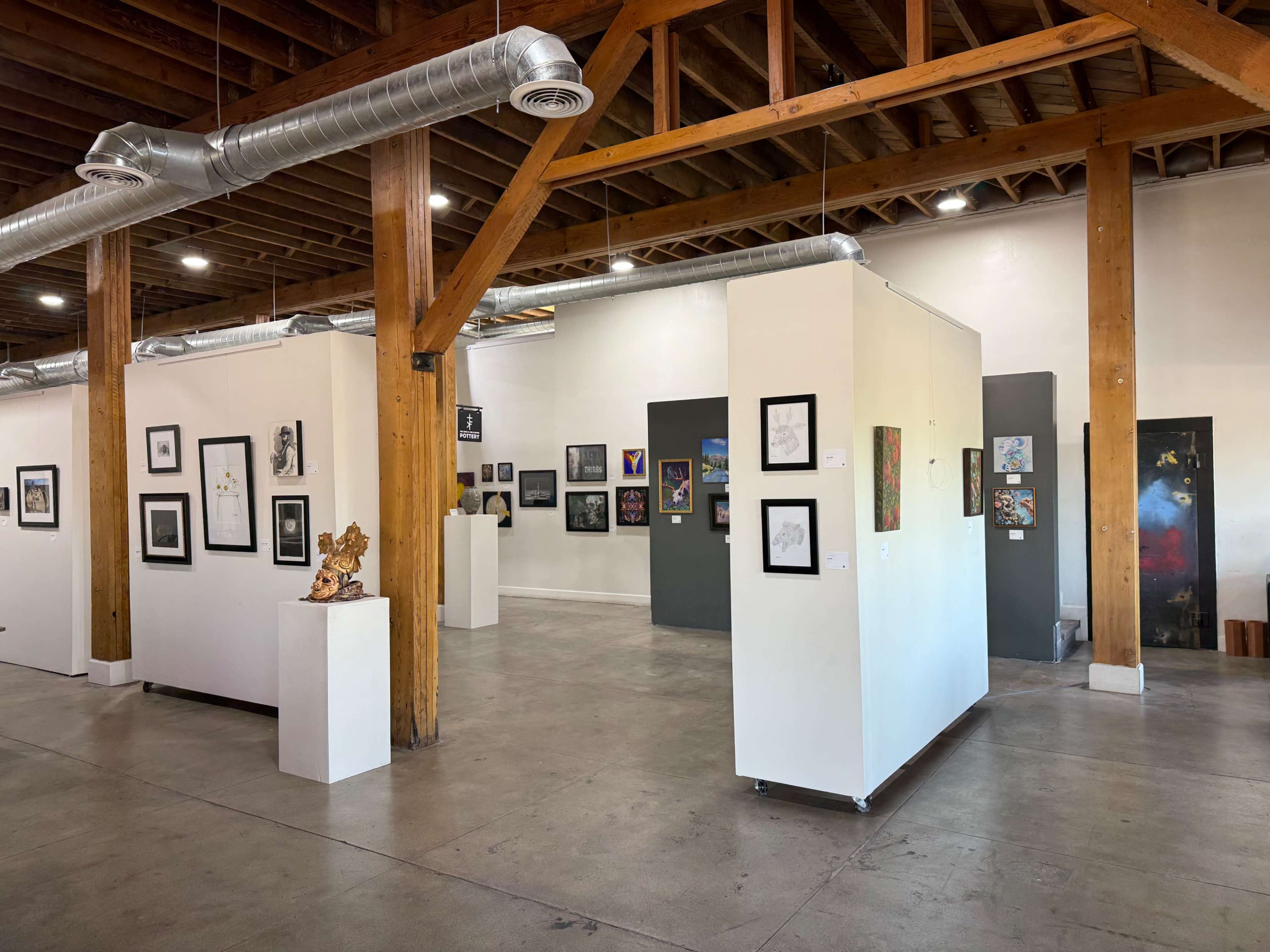 The image shows an art gallery with various framed artworks displayed on white walls, wooden beams, and gray partitions.