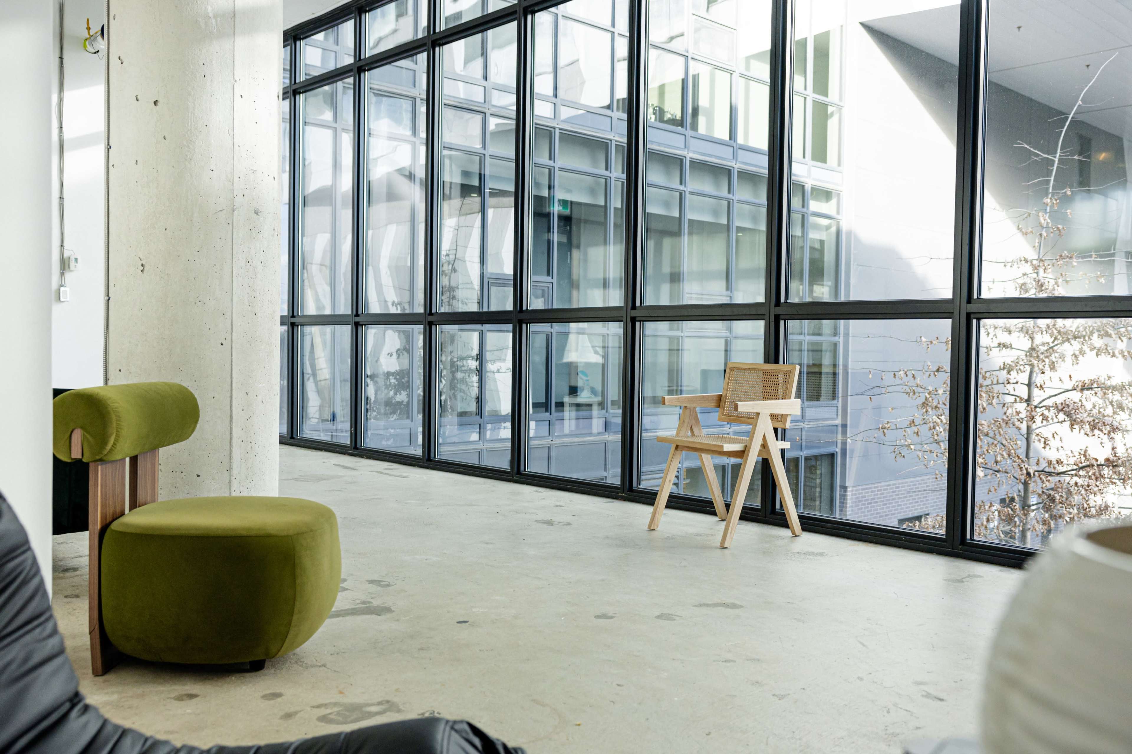 The image shows a minimalist interior space with large windows, a green upholstered chair, and a wooden chair positioned on a concrete floor.