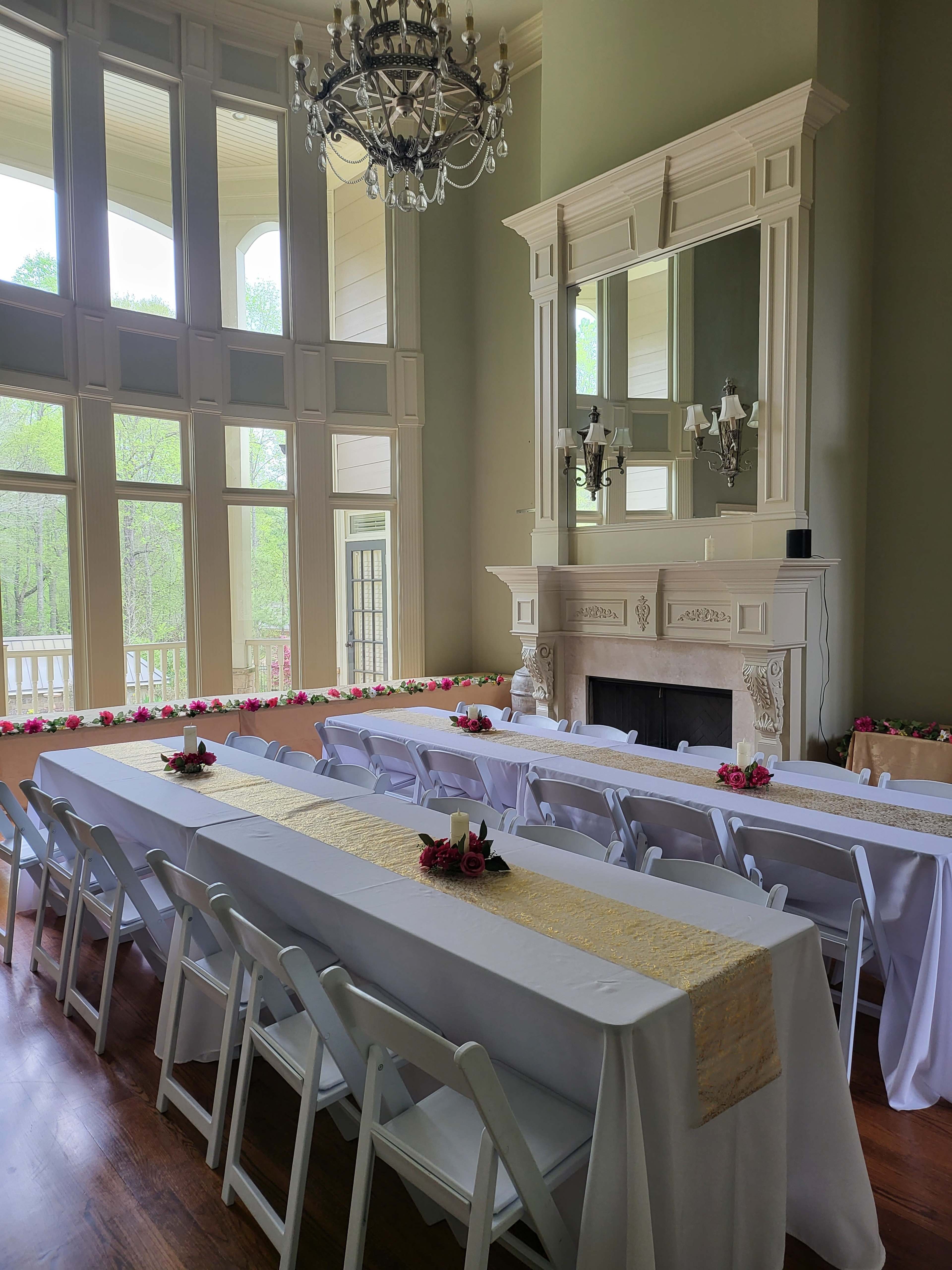A spacious room with large windows, featuring multiple long tables set with white tablecloths and decorative centerpieces, alongside a fireplace and a chandelier above.