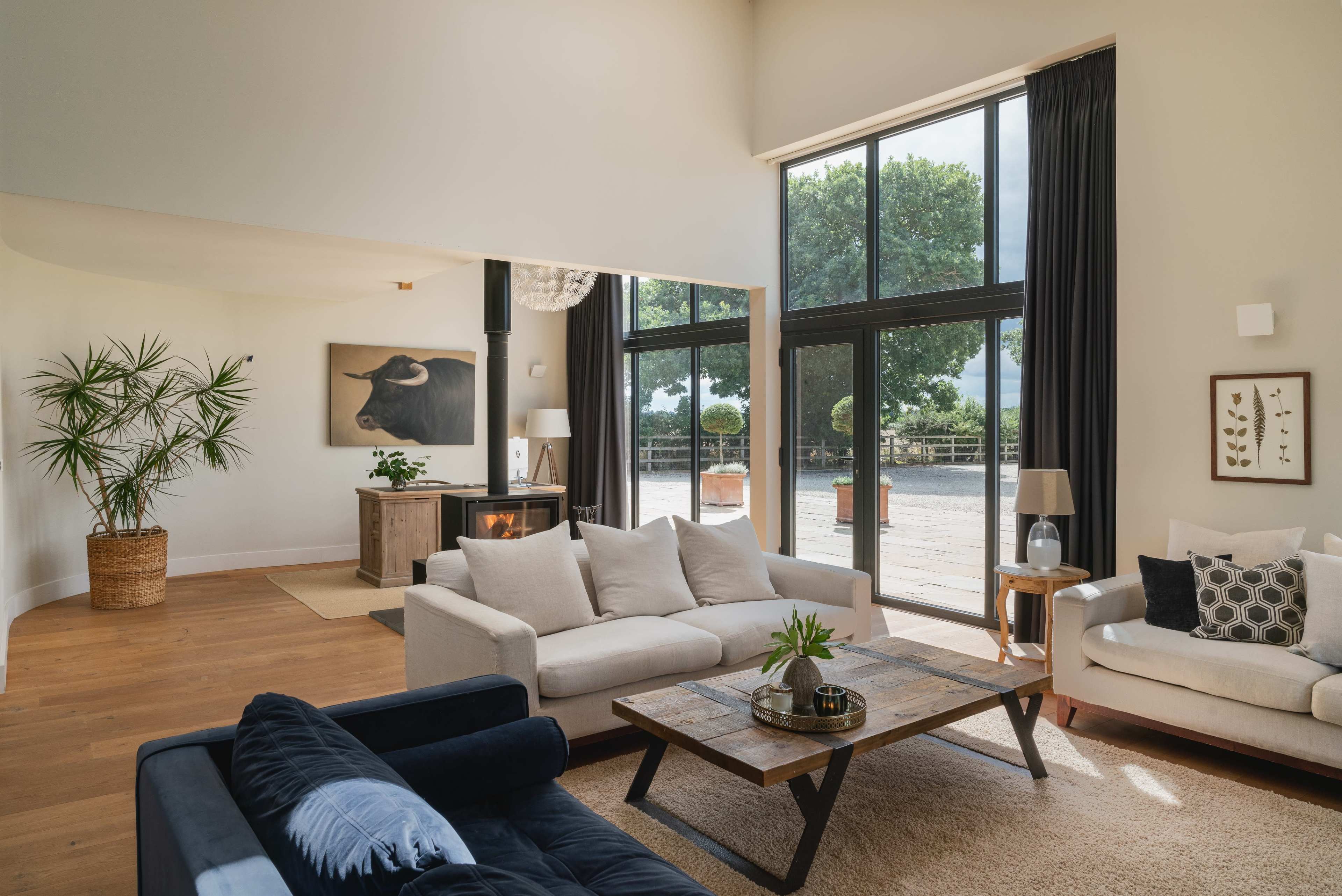 A spacious living room featuring a light-colored sofa, a dark blue couch, a wooden coffee table, and large windows with views of greenery outside.