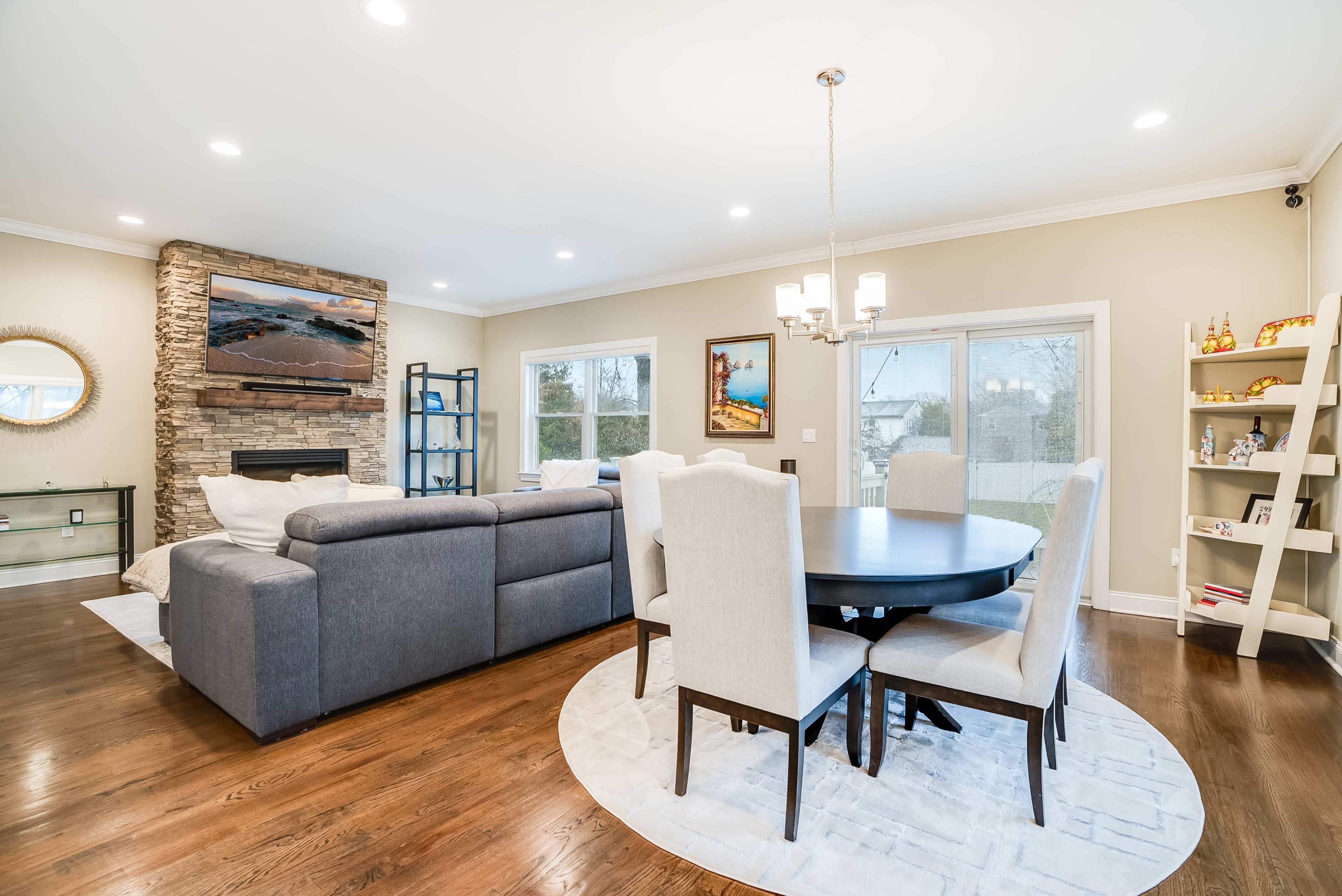 A modern living room with a circular dining table, upholstered chairs, a stone fireplace, and large windows allowing natural light.