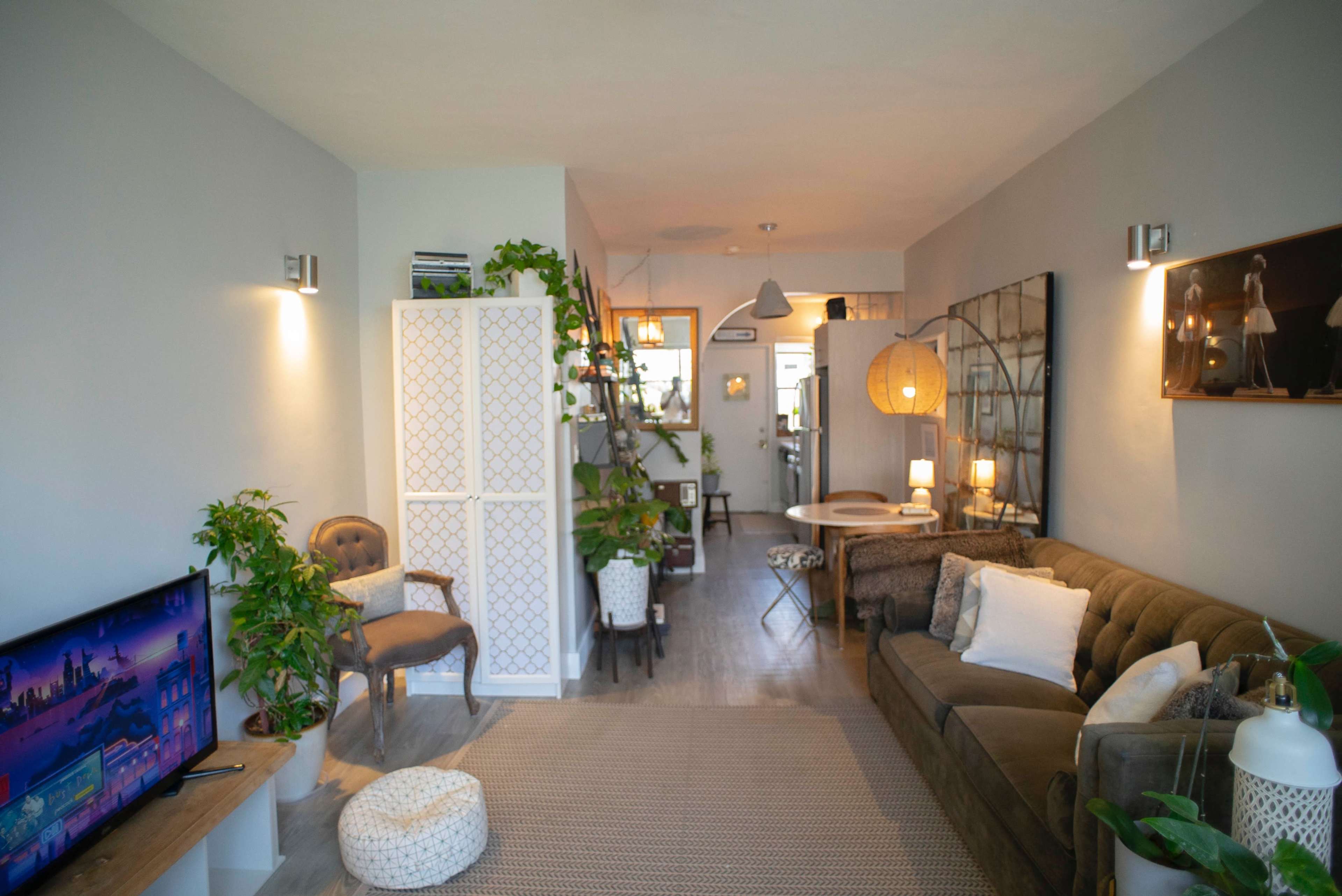 A cozy living room with a sofa, a television, plants, and a dining area in the background.