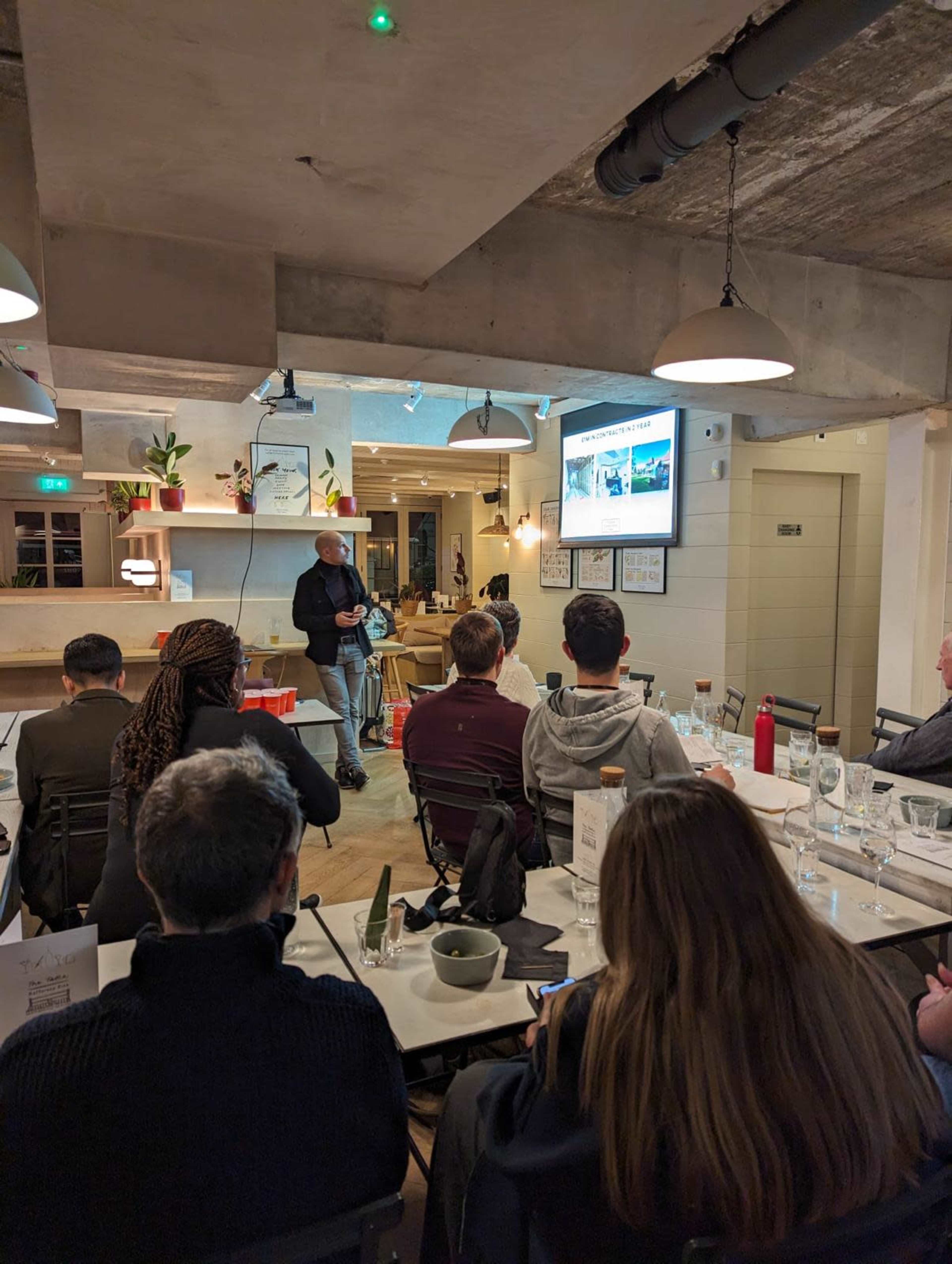 A speaker presents in a well-lit restaurant setting, with attendees seated at tables watching a screen displaying visuals.