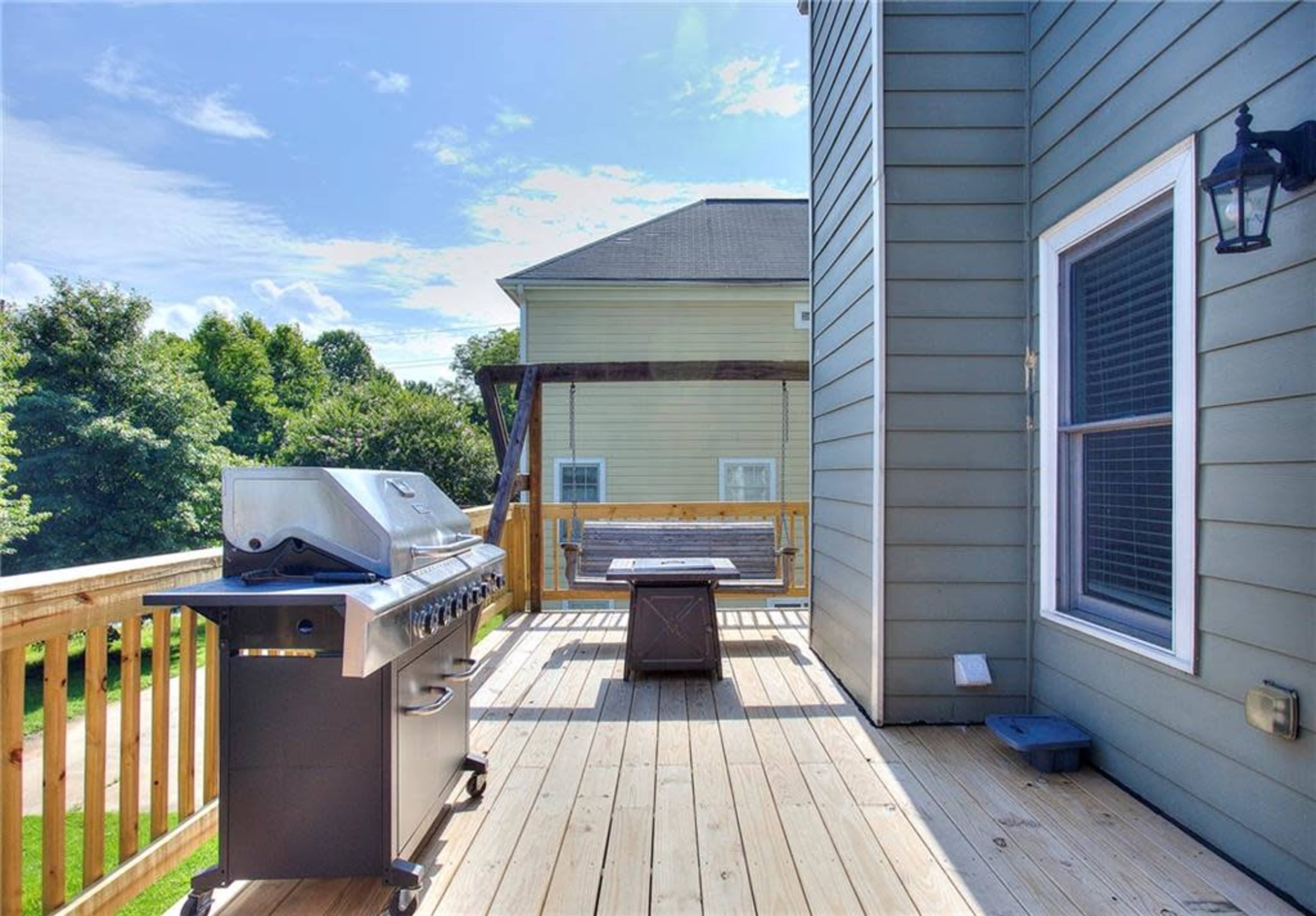 A wooden deck with a grill and a table, surrounded by greenery and residential buildings.