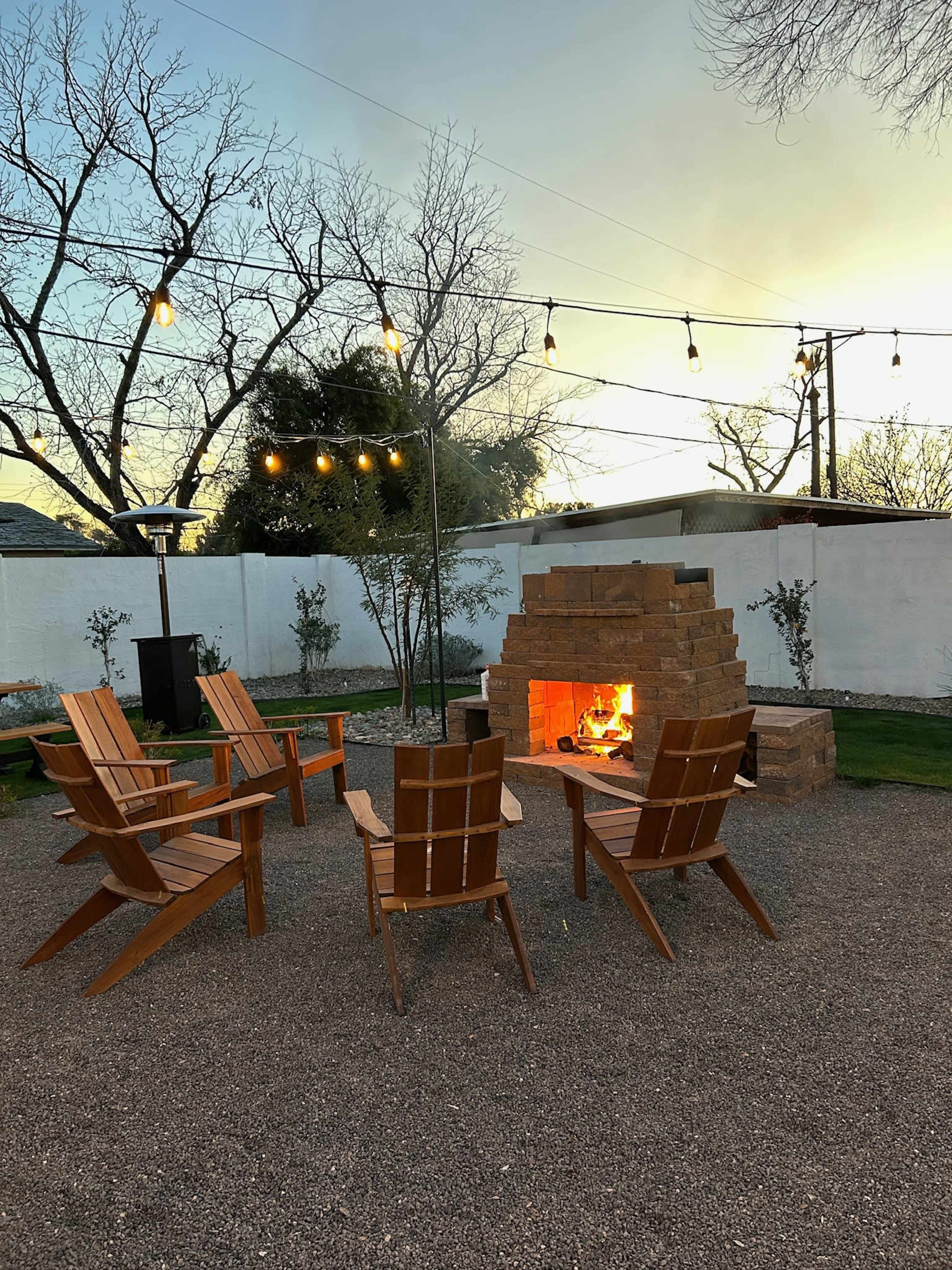 A circle of wooden chairs surrounds a stone fire pit, illuminated by string lights against a twilight sky.