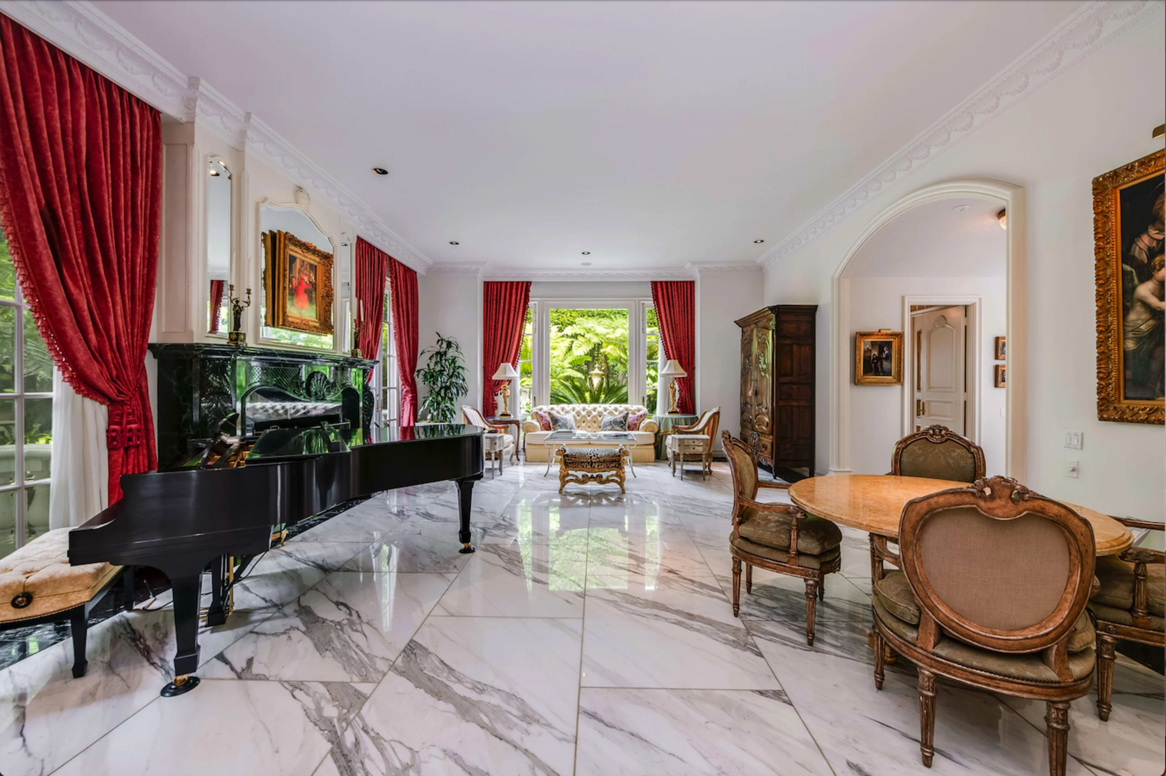The room features a grand piano, marble flooring, and large windows dressed with red curtains, allowing natural light to illuminate the space.