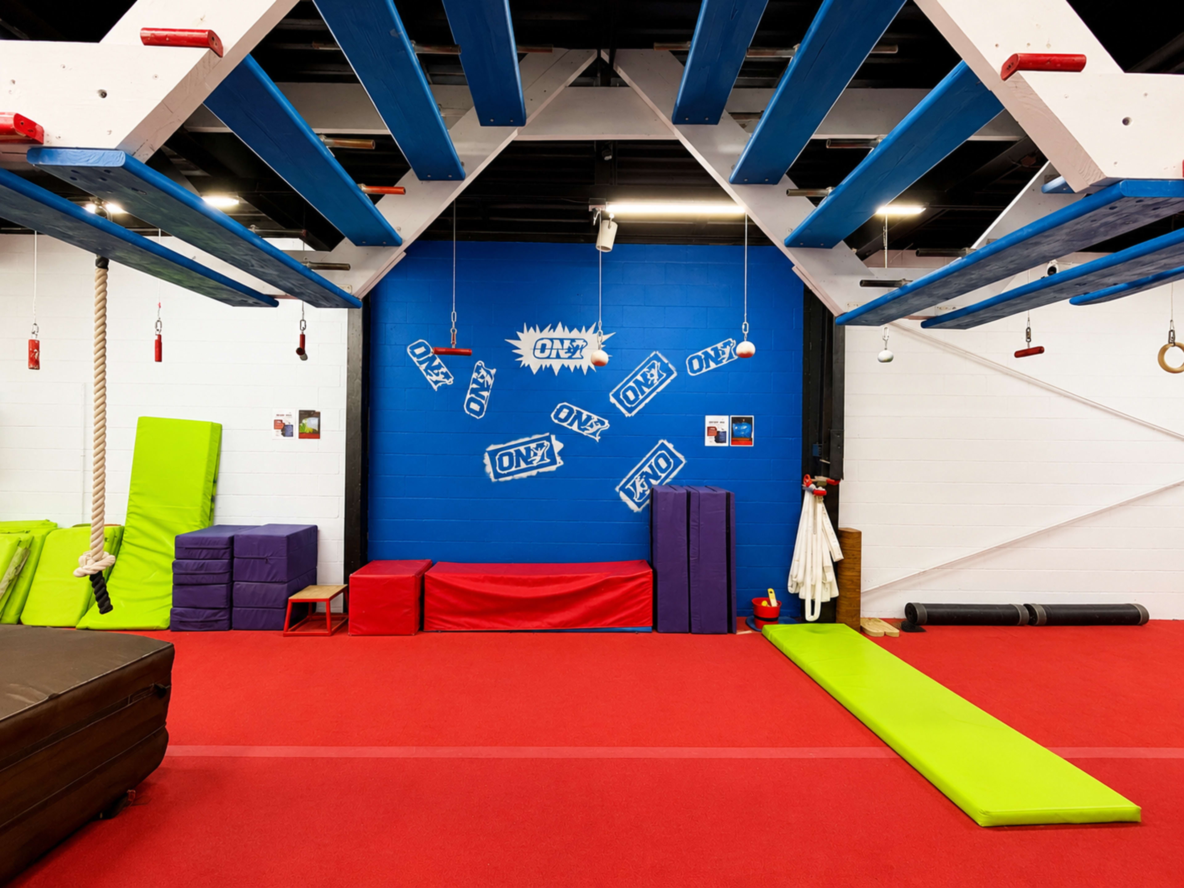 The image shows an indoor obstacle training facility with colorful mats, ropes, and a blue wall featuring various logos.