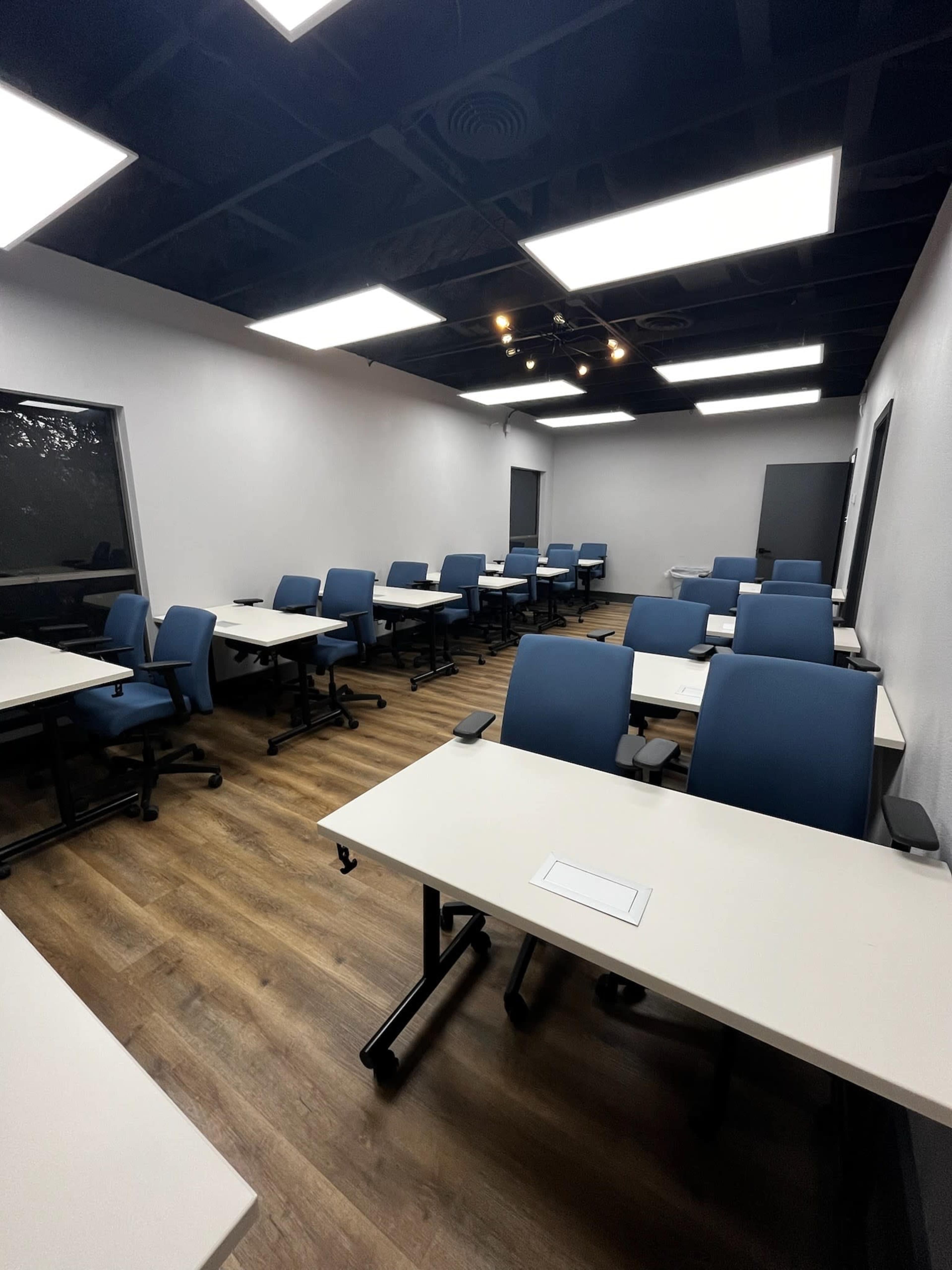 The image shows an empty classroom with multiple blue chairs and white tables arranged in a grid pattern under bright overhead lights.