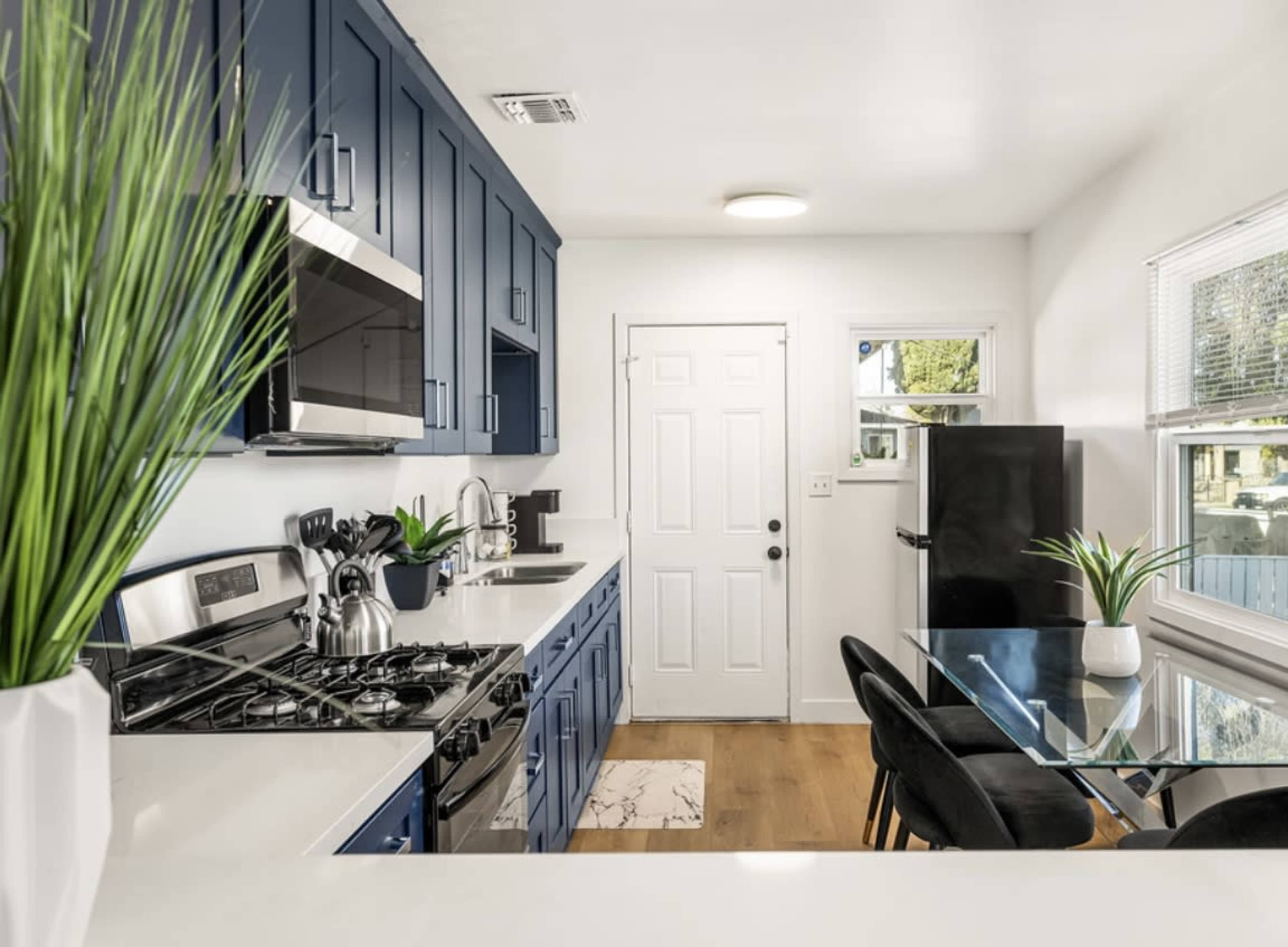 A modern kitchen with navy blue cabinets, a stainless steel gas stove, a glass dining table, and a door leading to the outside.