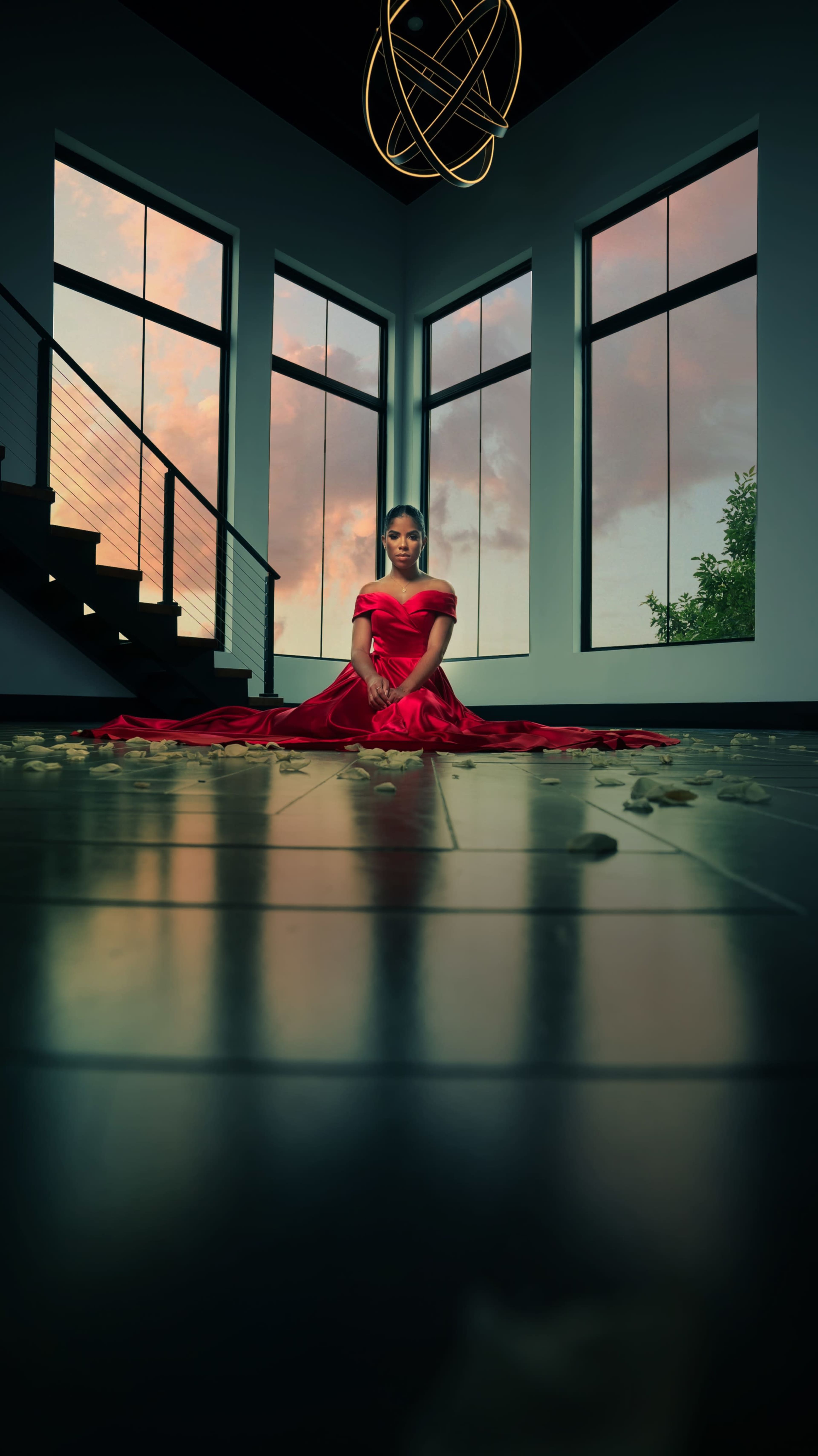 A woman in a red dress sits on the floor of a modern room with large windows, surrounded by scattered petals.