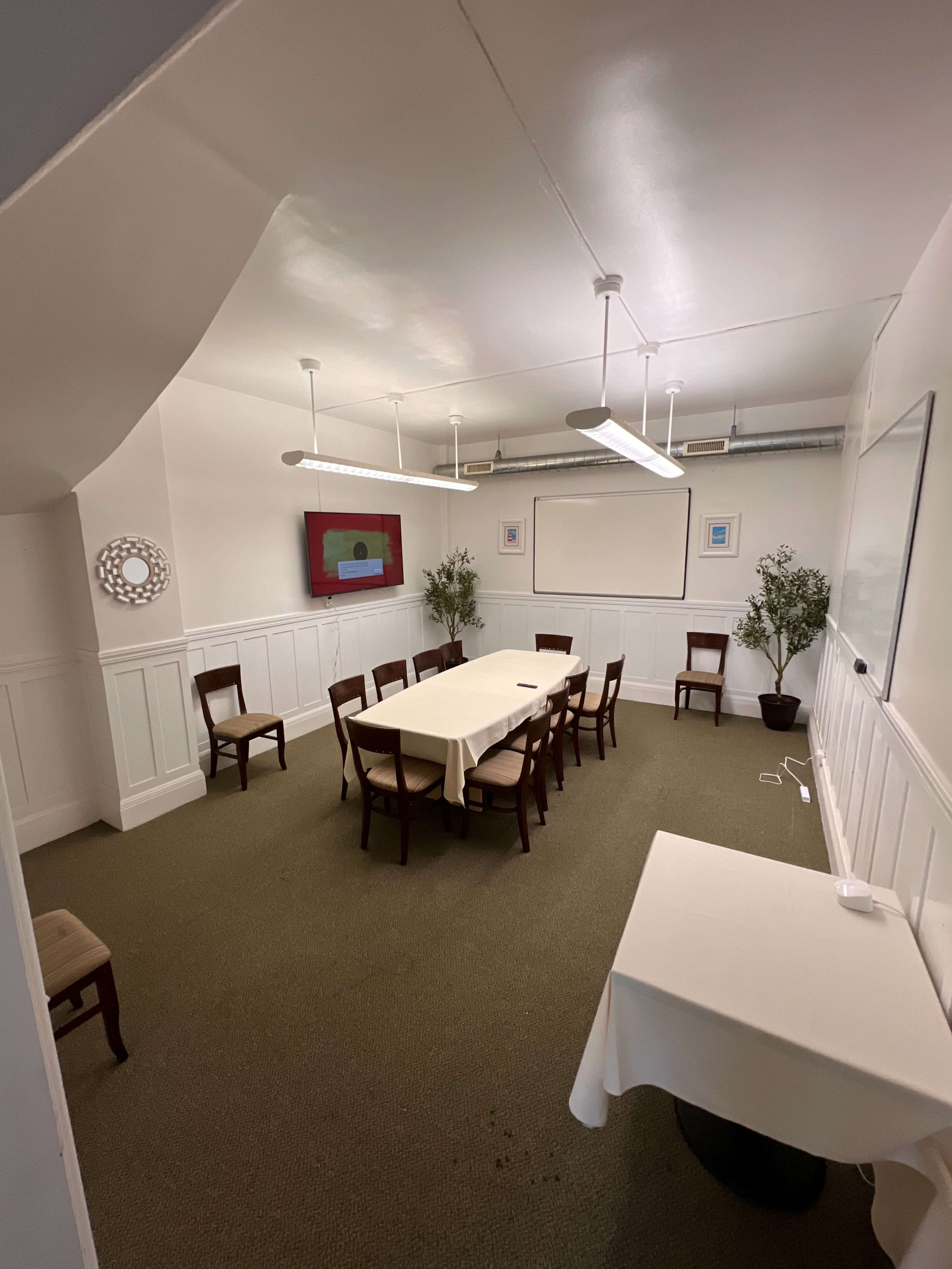 The image shows a sparsely furnished meeting room with a long table surrounded by chairs, plants in the corners, and a screen mounted on the wall.