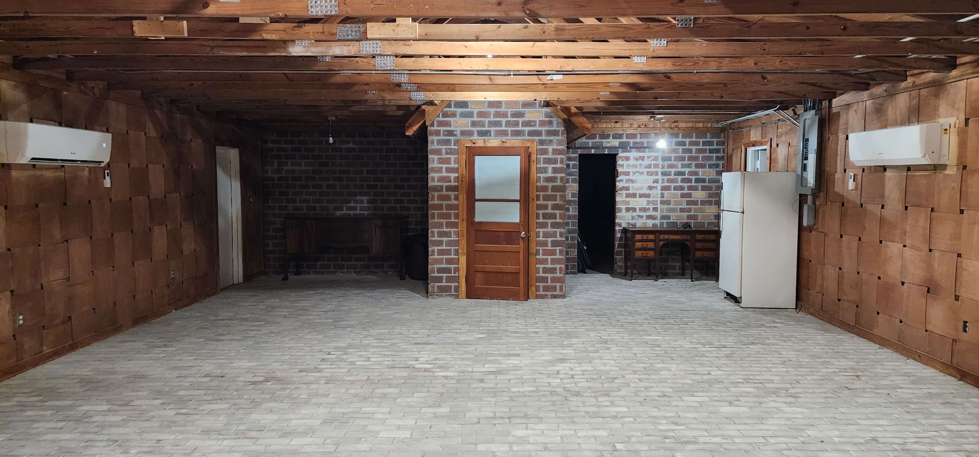 A spacious room with brick walls, wooden beams overhead, and a concrete floor, featuring a wooden door, a small table with chairs, and a white refrigerator against one side.