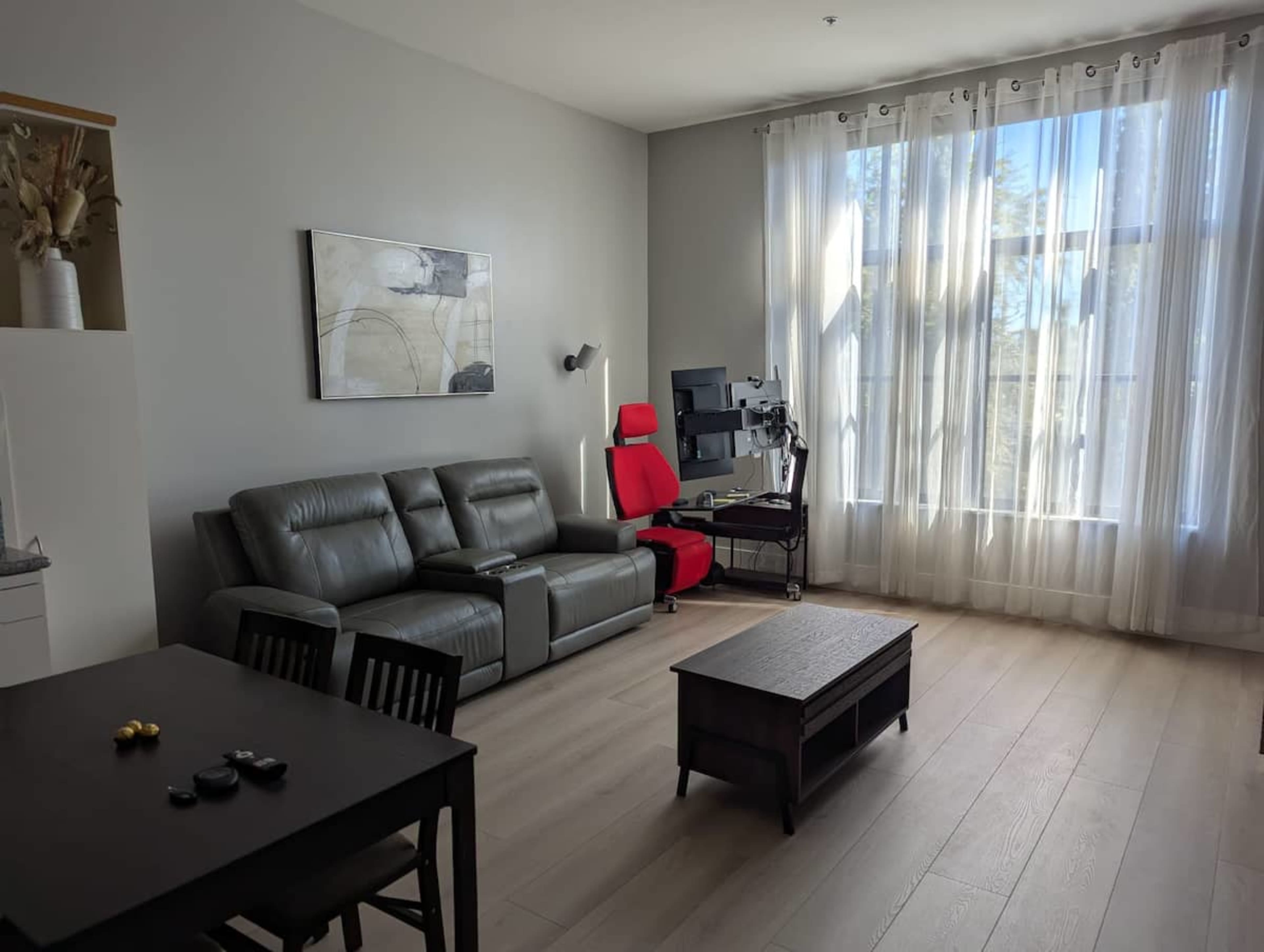The image shows a modern living room featuring a gray sofa, a black dining table with chairs, and a workspace with a computer setup near a large window with sheer curtains.