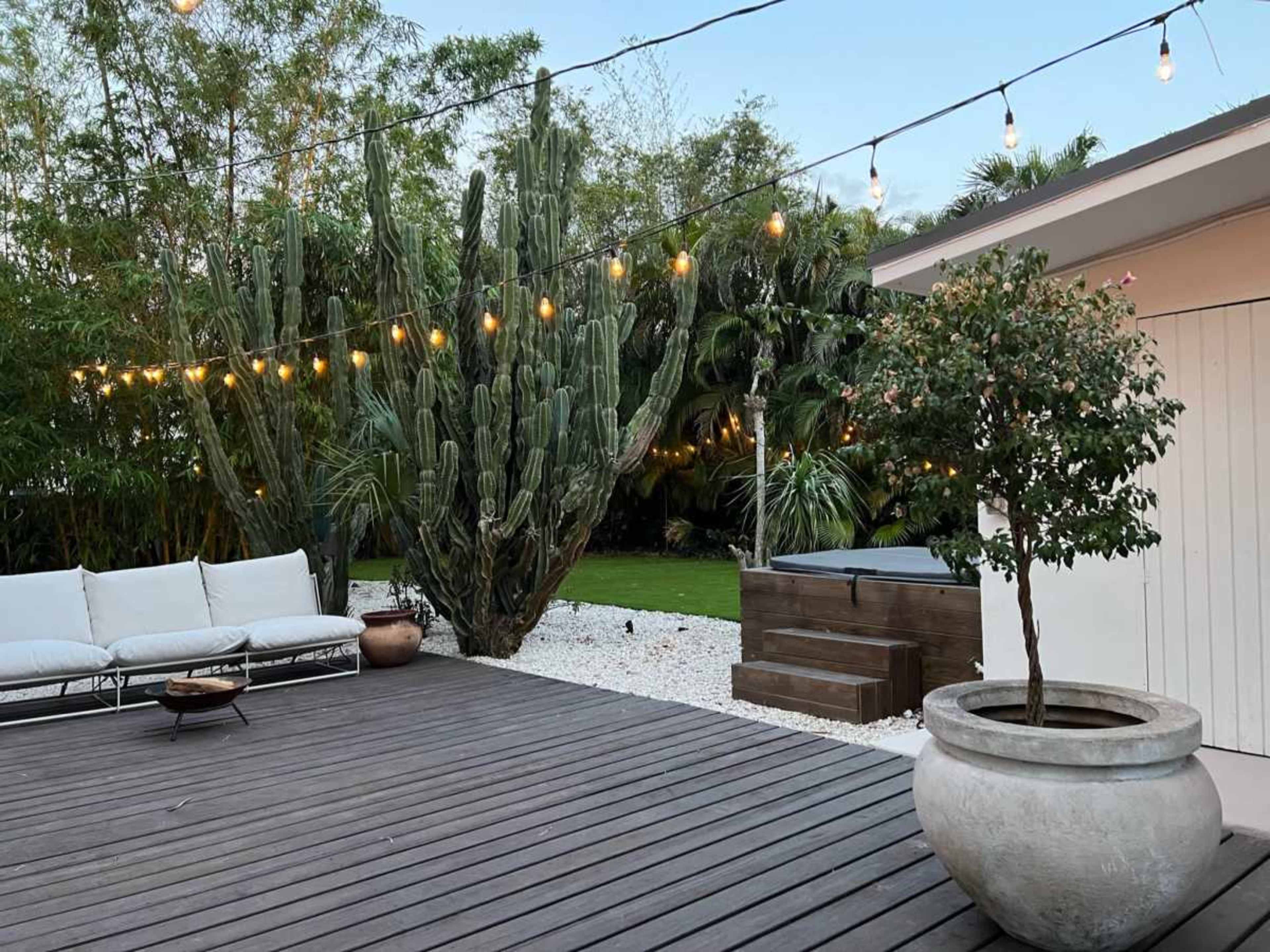 The image shows a wooden deck area with outdoor seating and string lights above, surrounded by lush greenery and cacti.