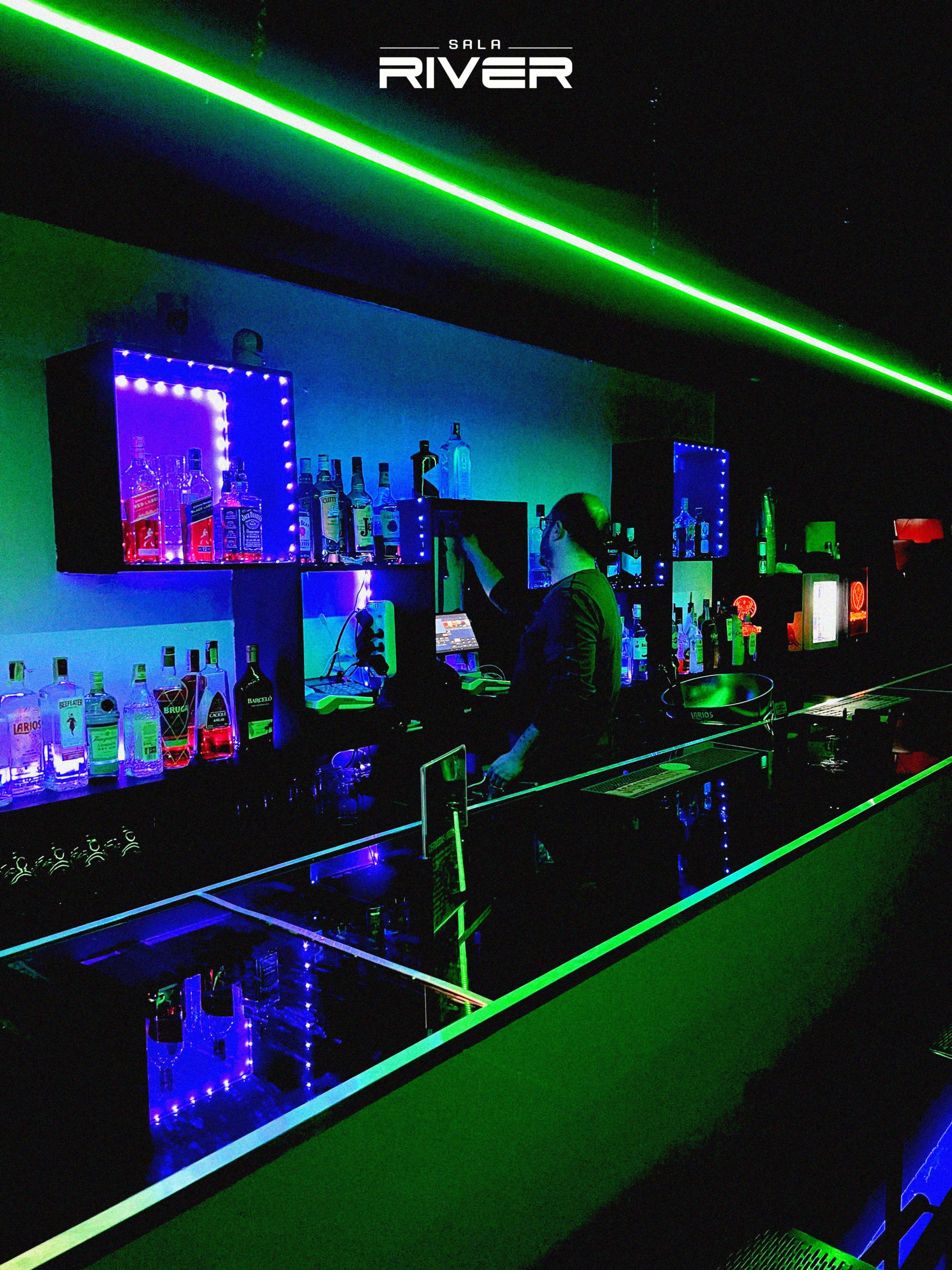 A bartender prepares drinks behind a sleek bar illuminated by green and blue LED lights, with various bottles displayed on shelves.
