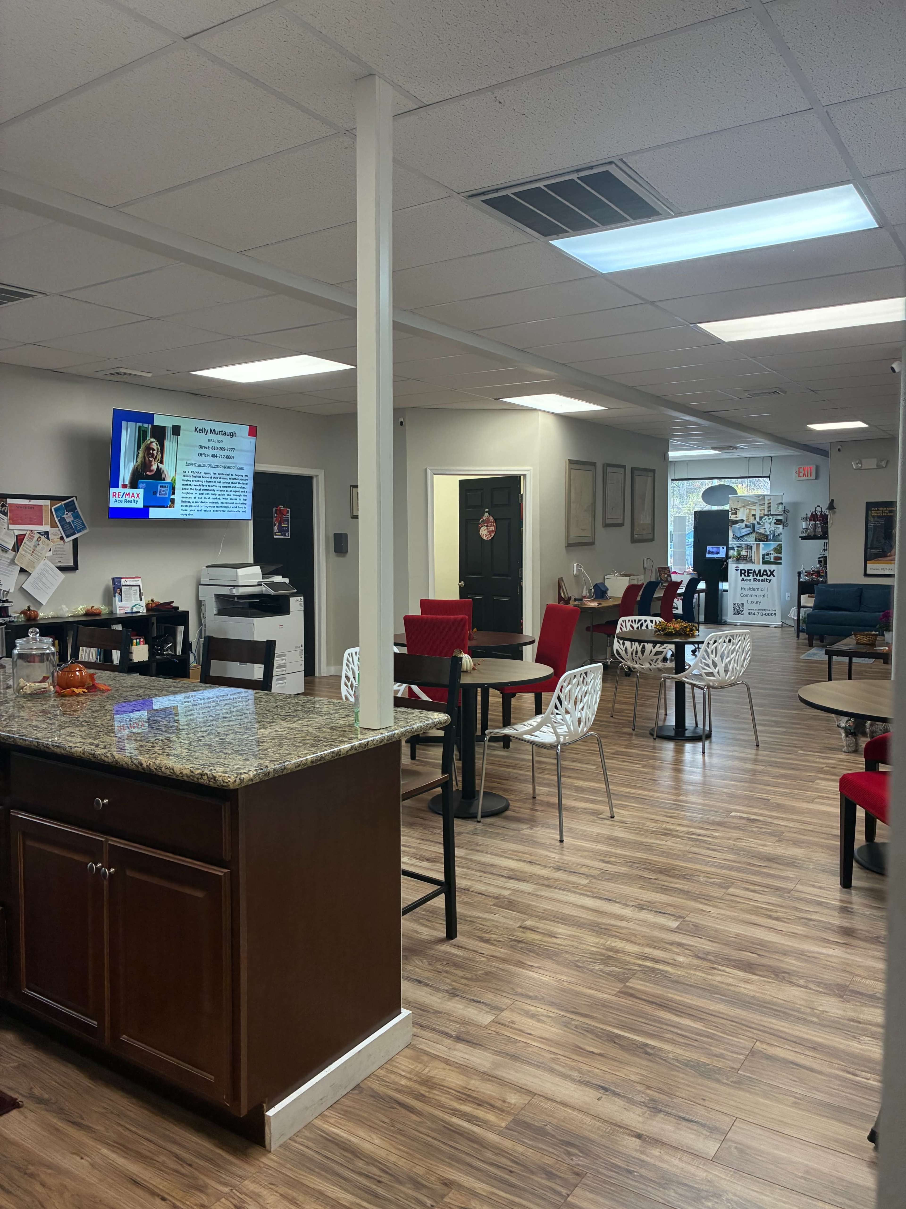 The image shows a modern office or waiting area with a reception desk, several tables and chairs, and a television displaying information on the wall.