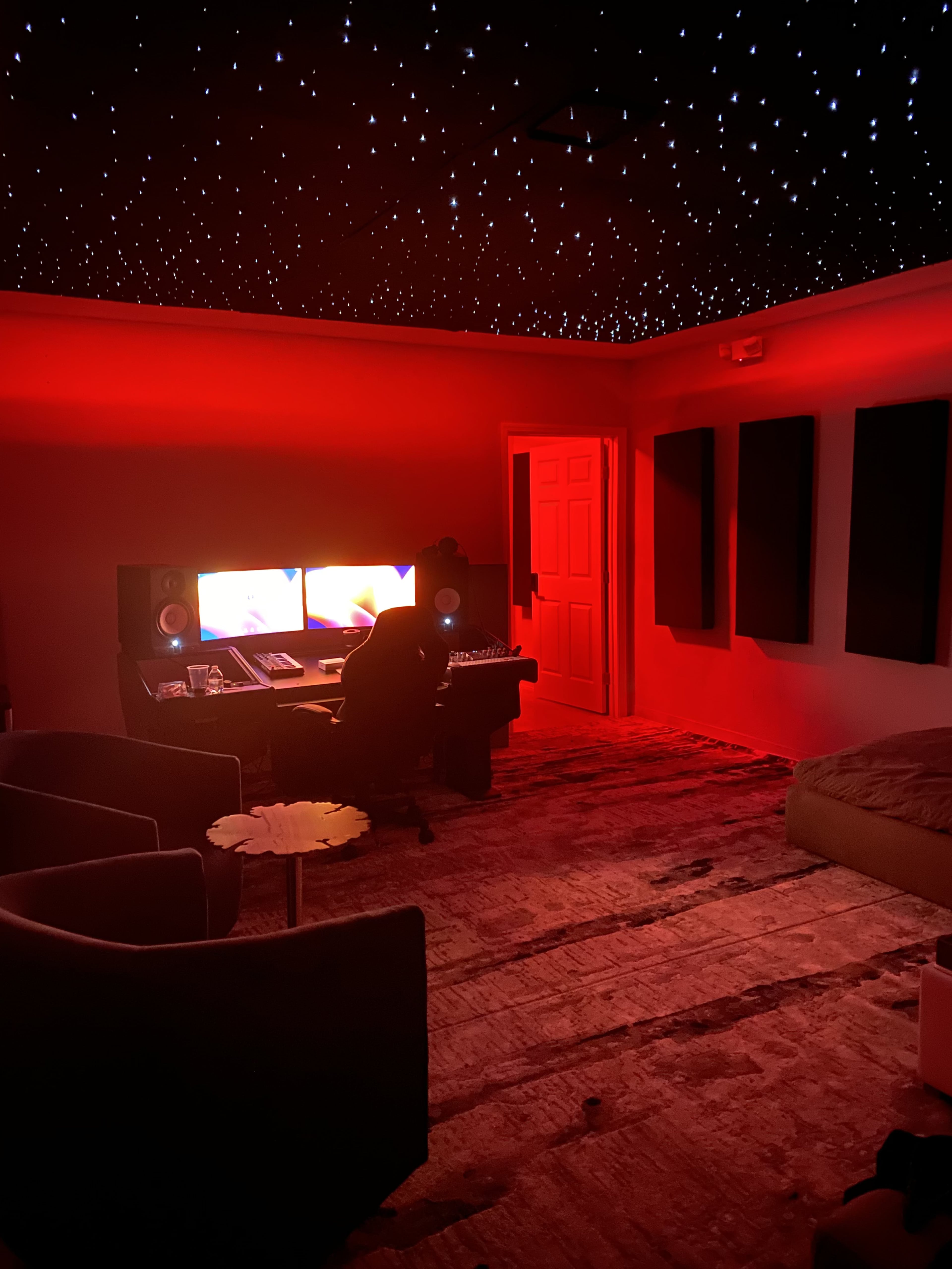 A dimly lit music studio features red lighting, a starry ceiling, and a central workstation with dual monitors.