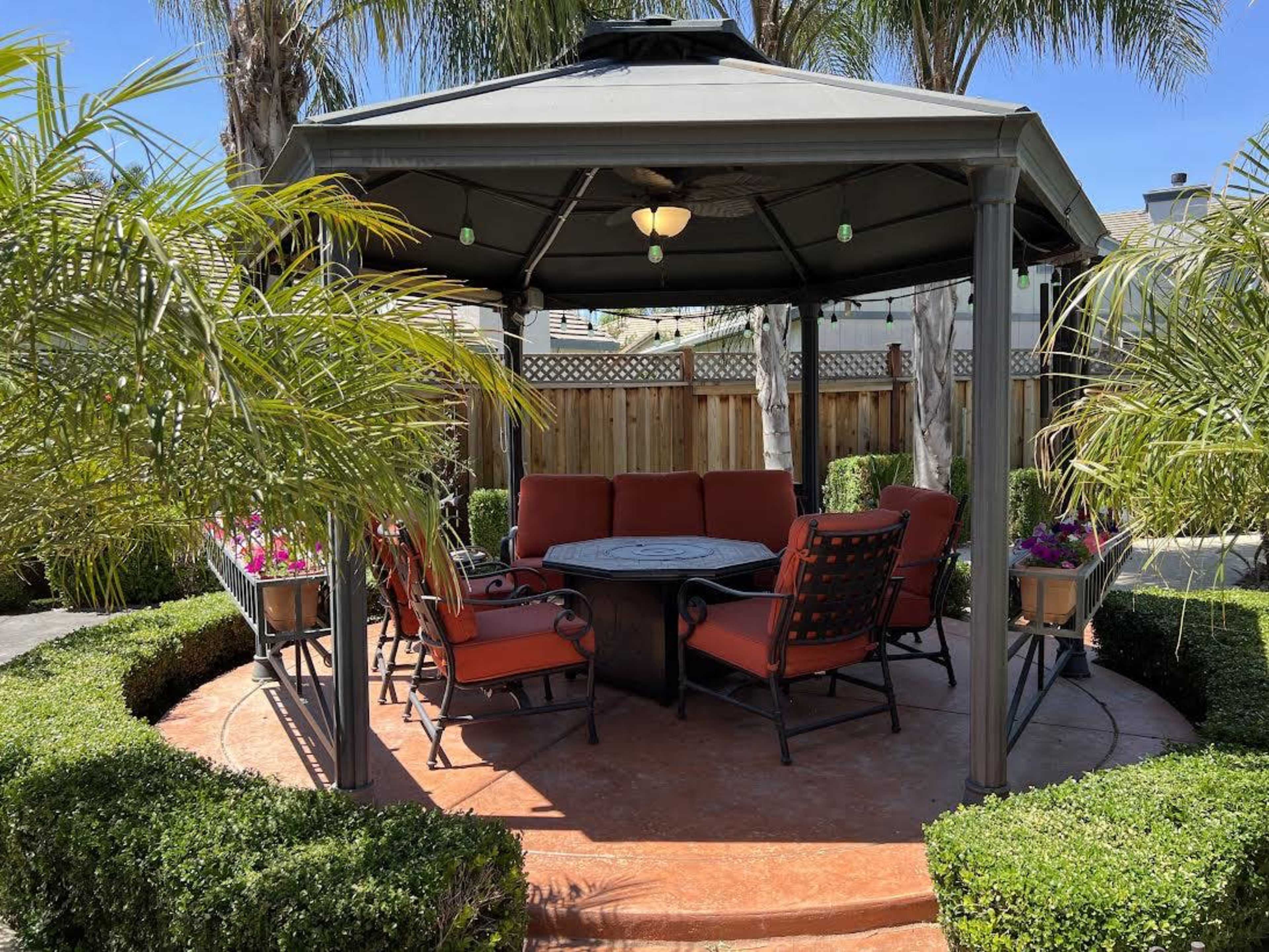 A gazebo with red seating surrounds a central table, set among neatly trimmed hedges and tropical plants.