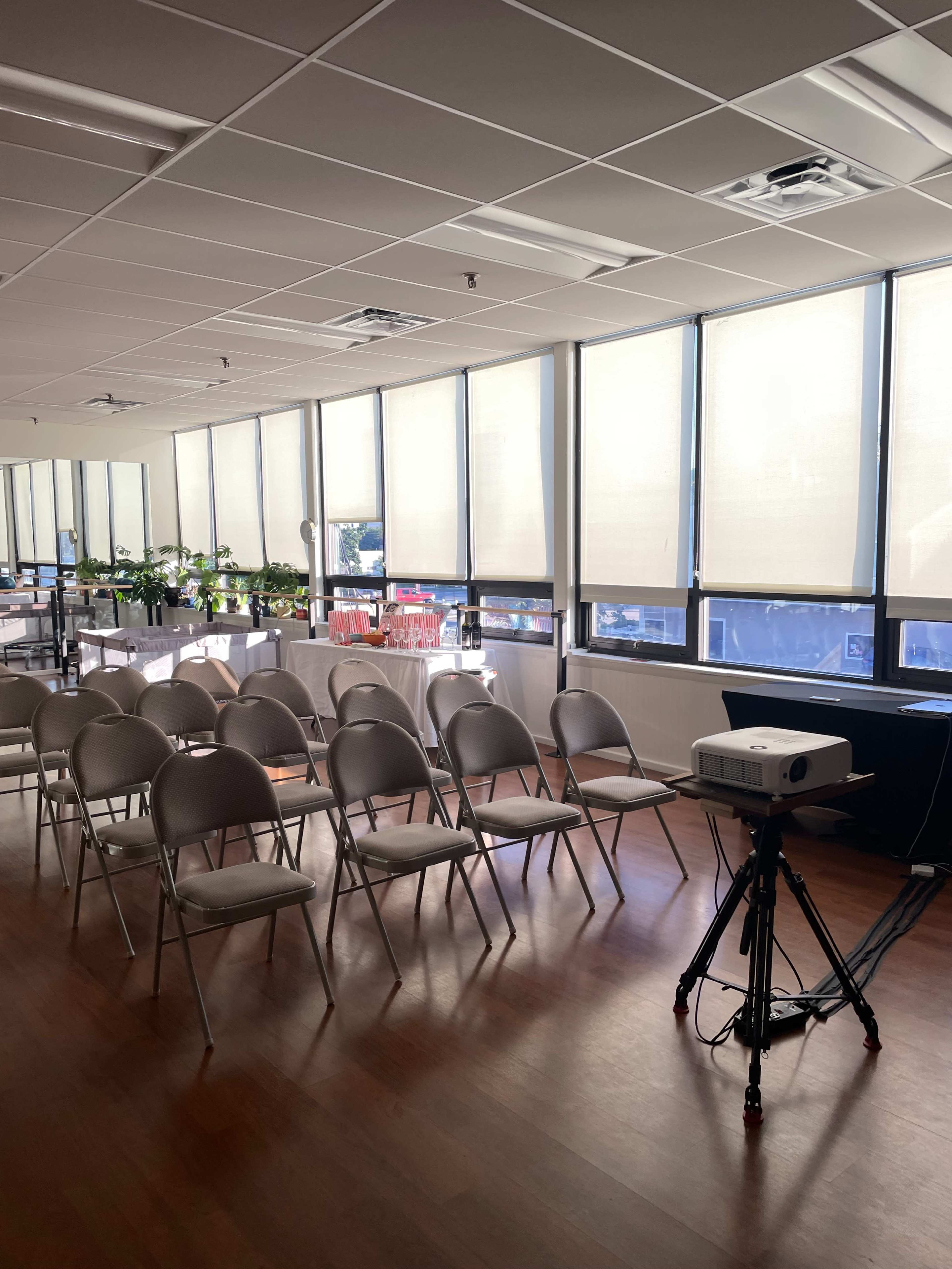 The image shows a spacious room set up for a presentation, featuring rows of folding chairs facing a projector on a tripod and large windows with natural light.
