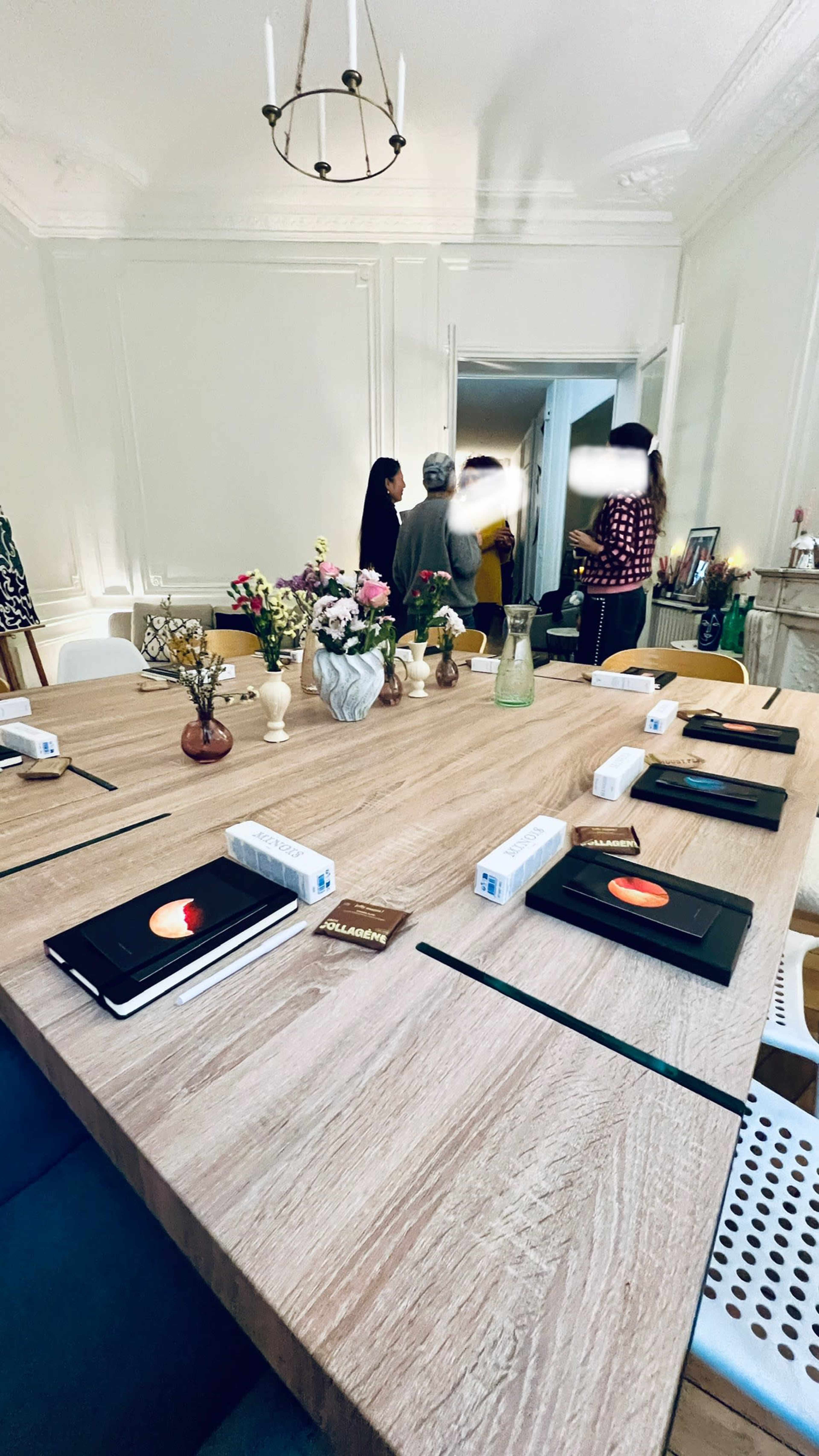 A large wooden table is set with notebooks and flowers in a bright room where people are gathered near a doorway.