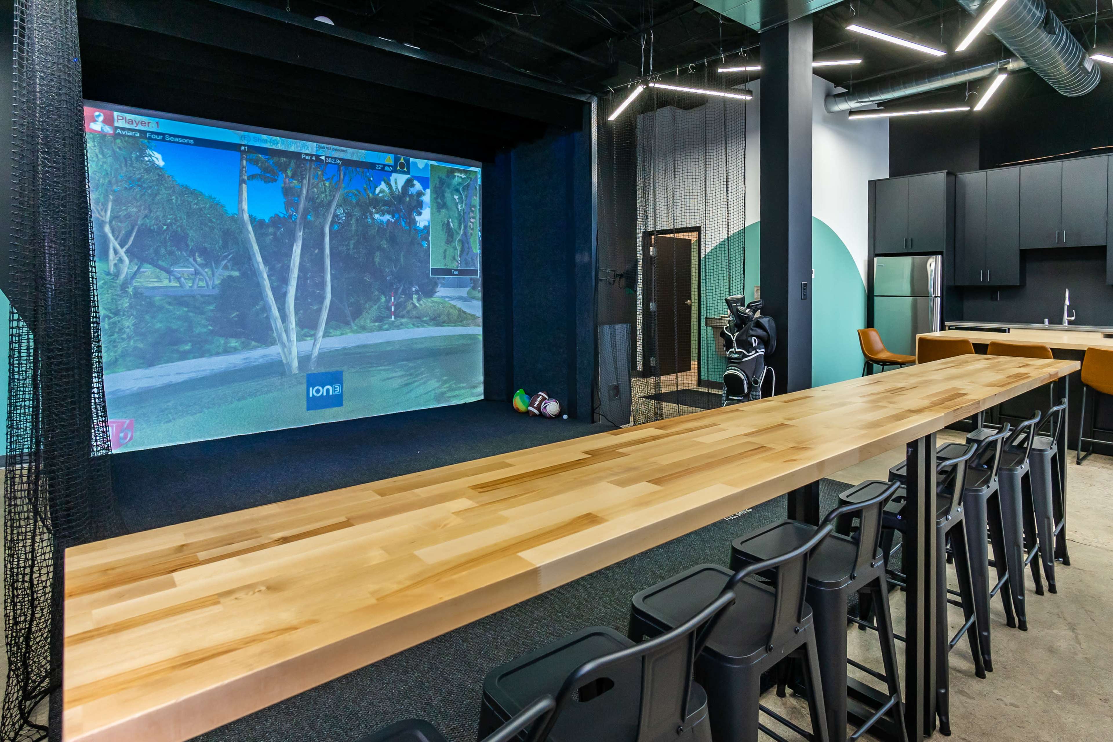A modern indoor golf simulation room featuring a large projection screen, a wooden table with bar stools, and a kitchenette in the background.